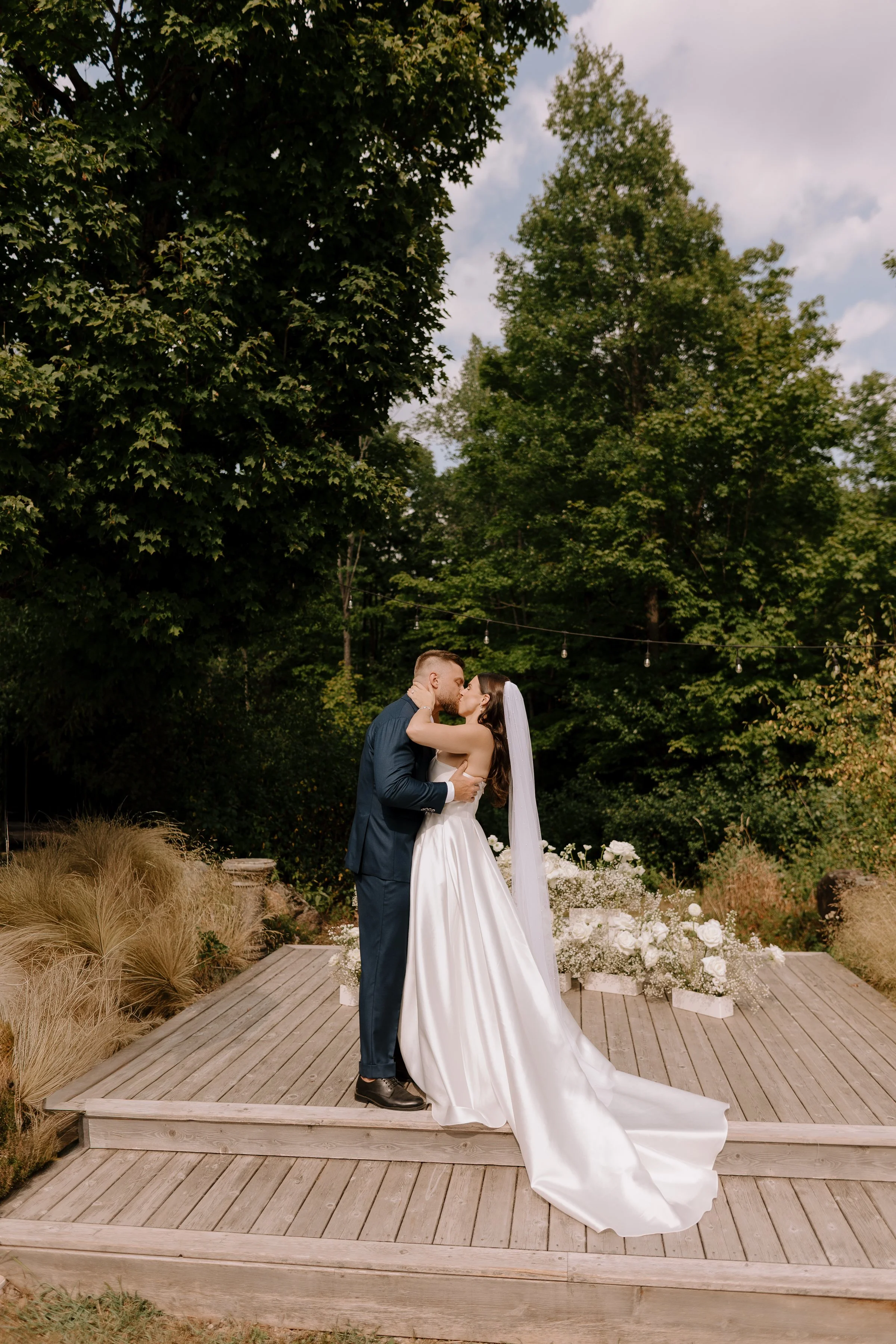 A bride and groom sharing a kiss on a wooden platform in an outdoor garden setting, surrounded by green trees and decorative plants.