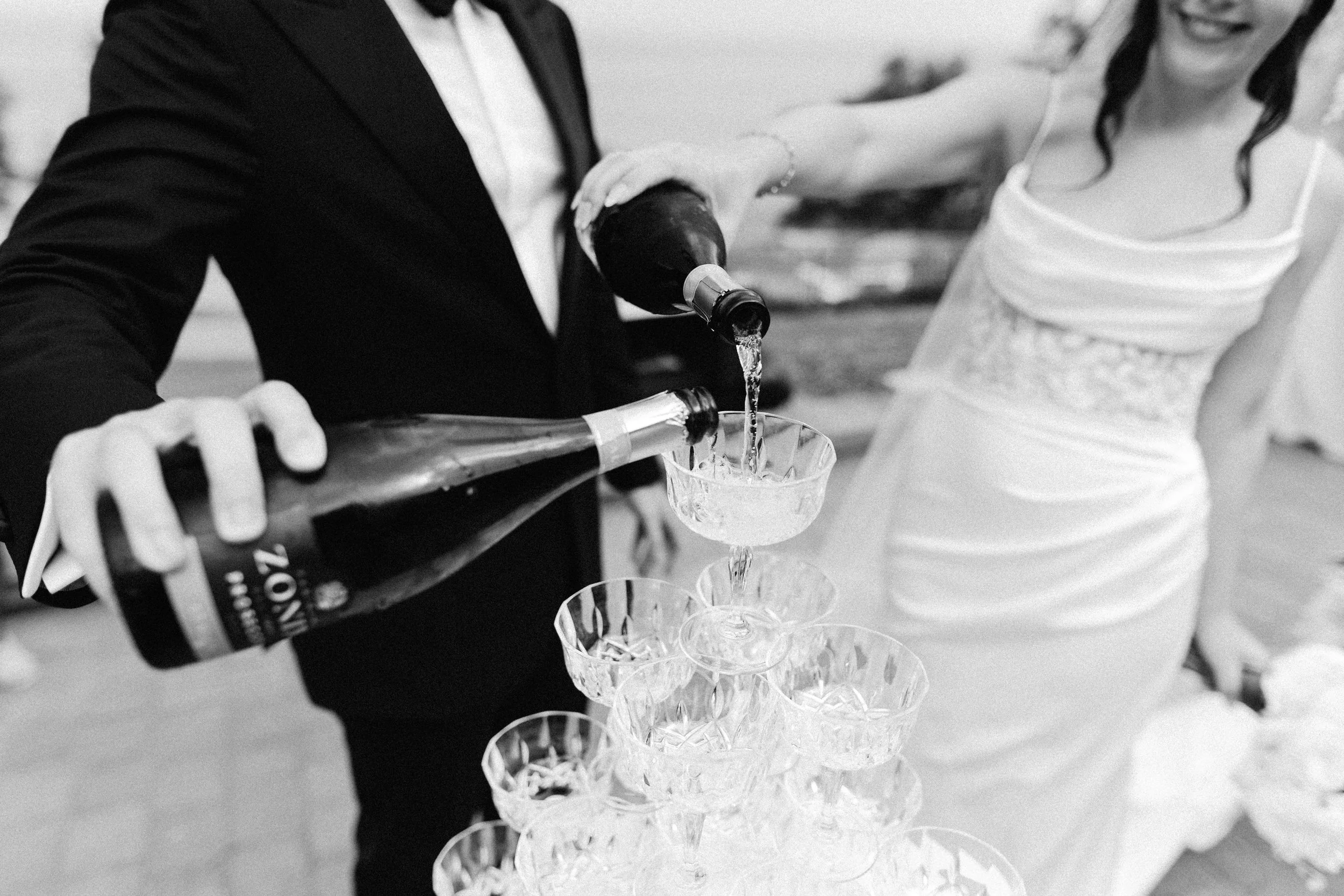 A person dressed in a suit is pouring champagne into a tower of glasses at a celebration or wedding.