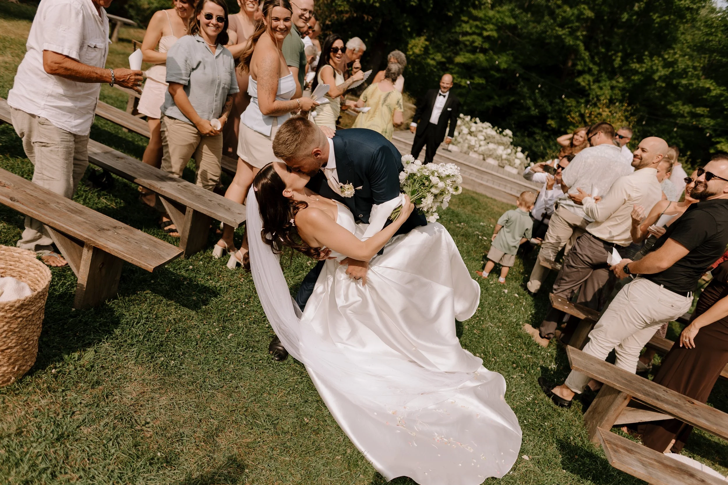 A bride and groom sharing a kiss during an outdoor wedding ceremony with guests standing and sitting on wooden benches, trees, and green grass in the background.