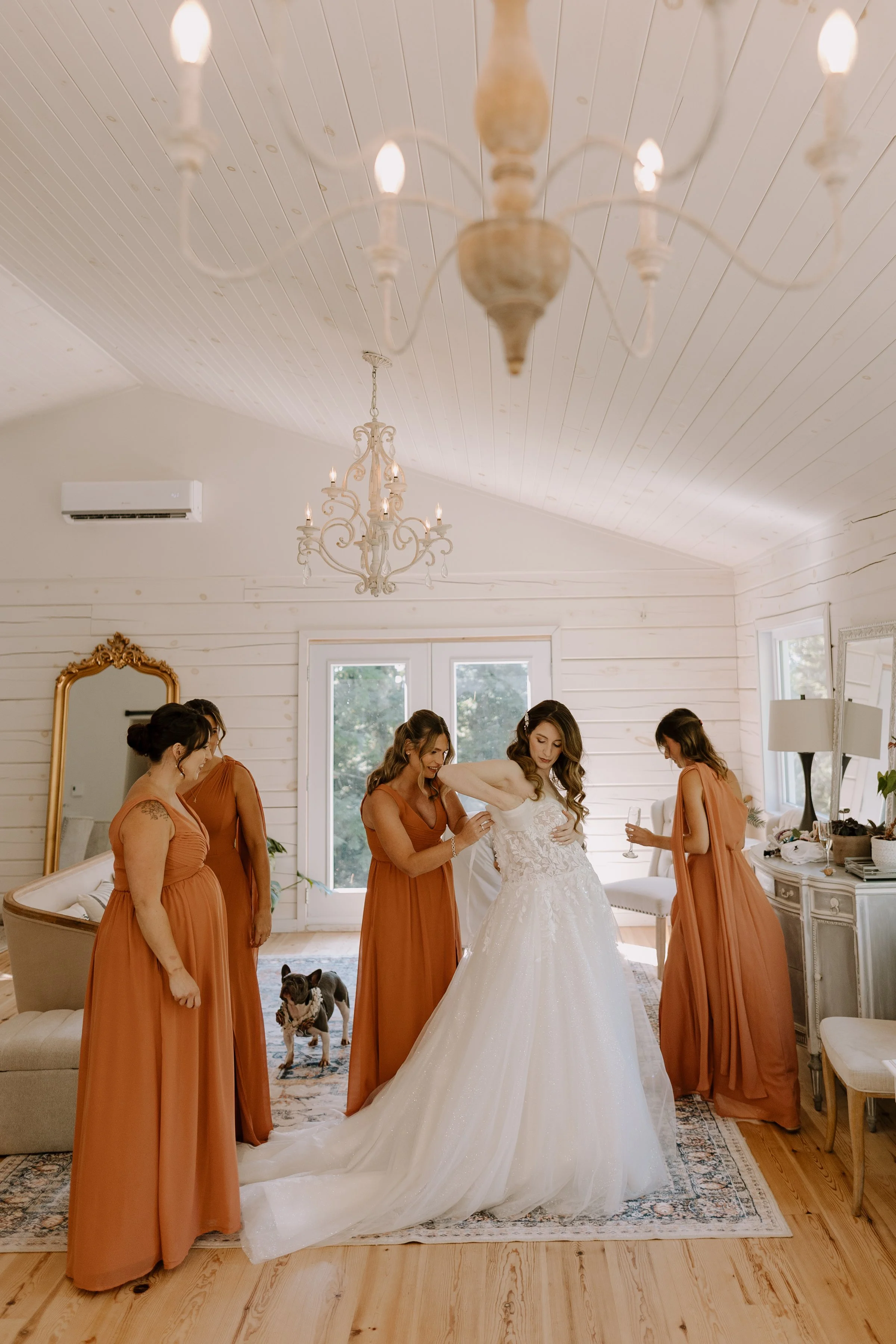 A bride in a white wedding gown surrounded by bridesmaids in long rust-colored dresses. The bride is helping to put on her gown, and the bridesmaids are standing nearby. The room has white walls, a chandelier hanging from the ceiling, and a French Bu