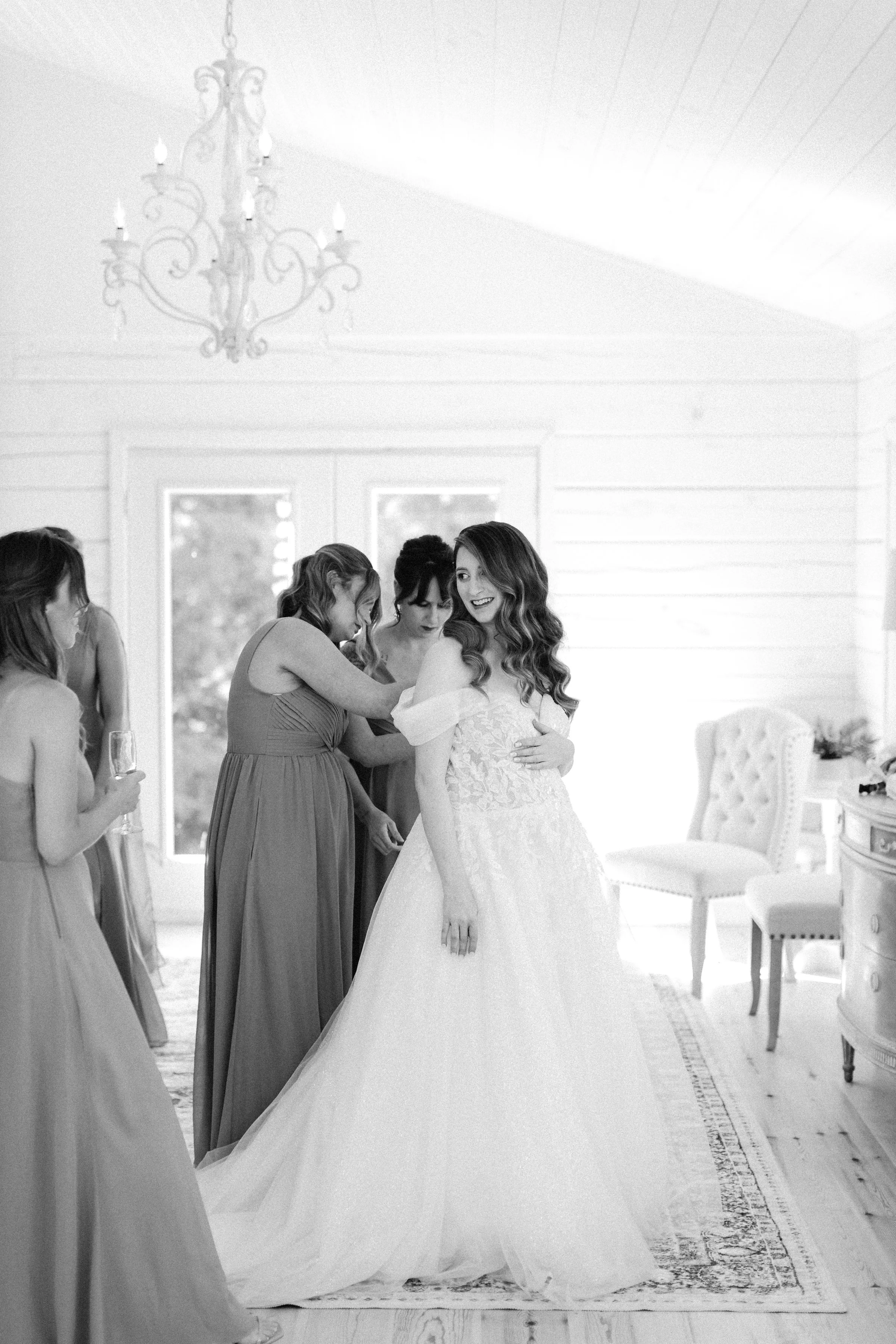 A bride is smiling while surrounded by bridesmaids in a bright room with large windows, a chandelier, and elegant furniture.