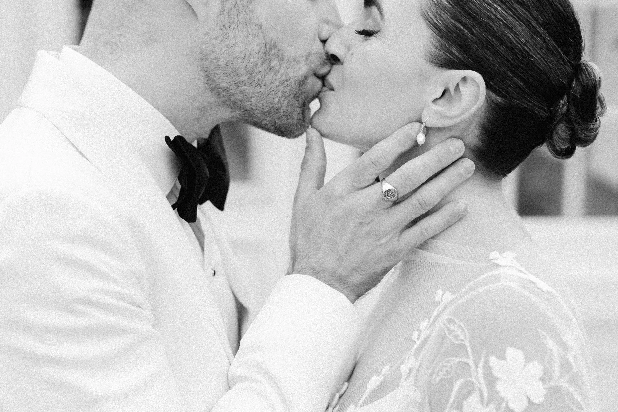 Black and white photo of a couple kissing, with the woman's hand gently touching the man's face. The woman is wearing a pearl earring and a ring, while the man is dressed in a tuxedo with a bow tie.