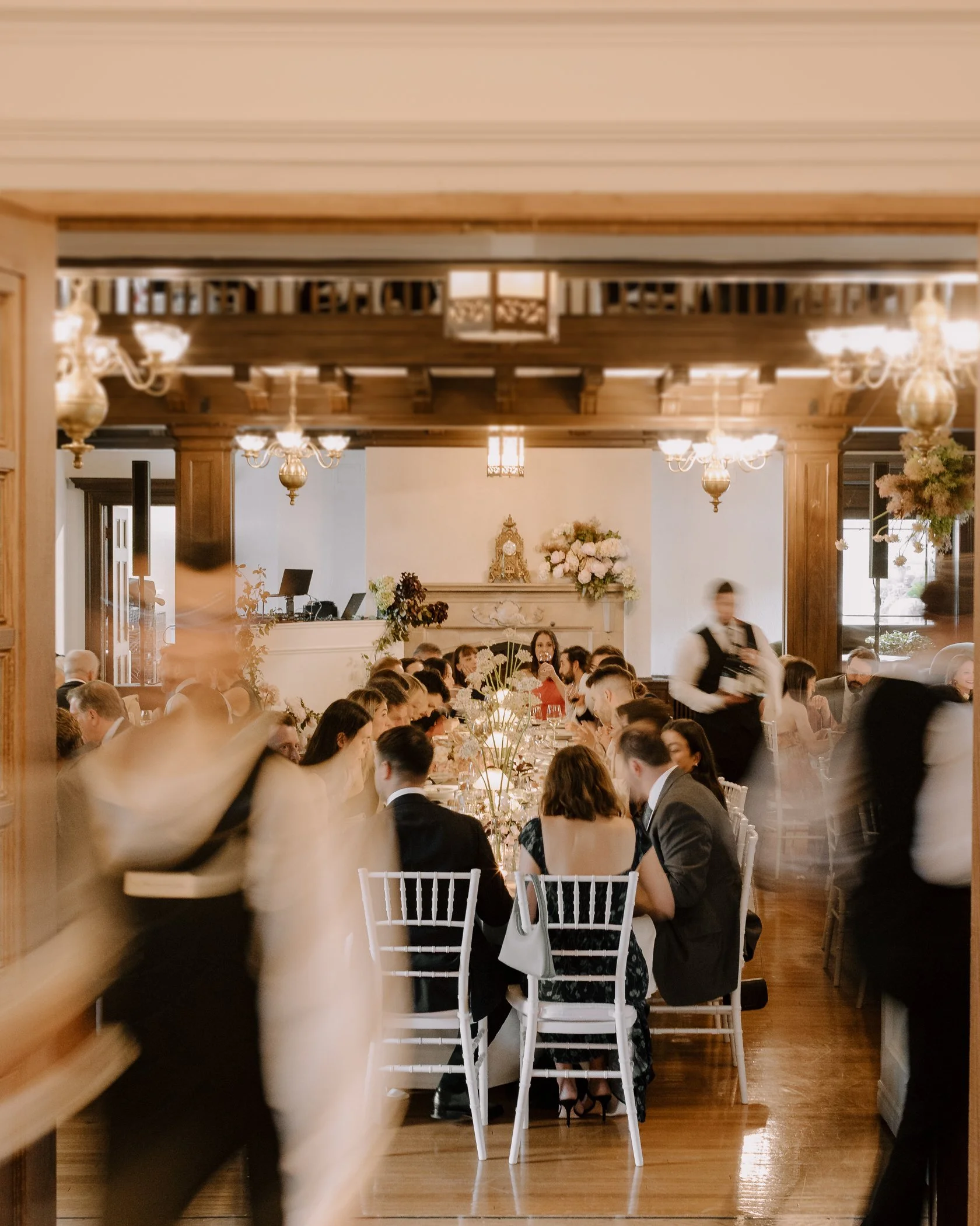 A busy indoor event with people seated at long tables, floral arrangements, warm lighting, wooden ceiling beams, and elegant chandeliers.