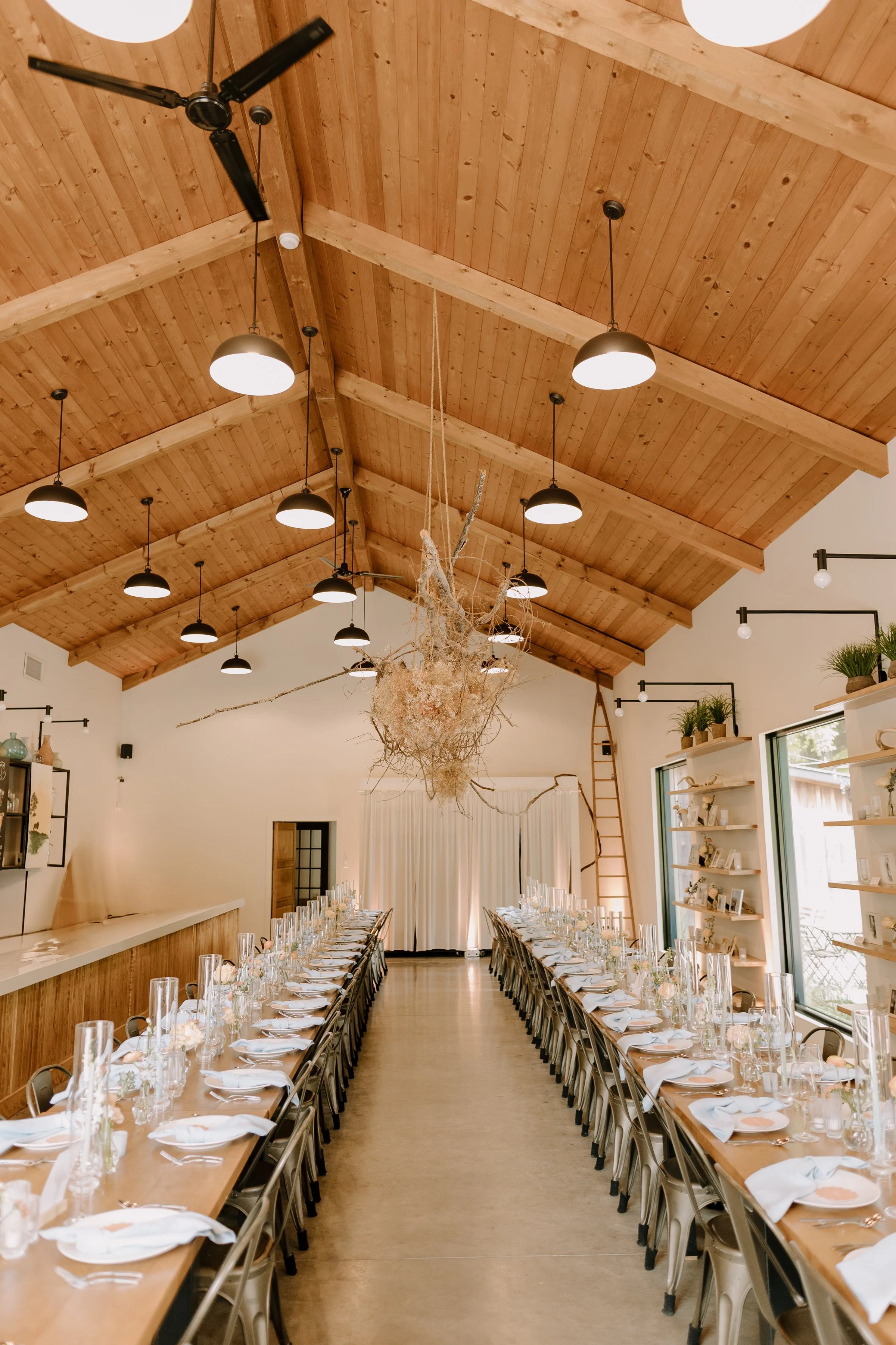 An indoor event space with a high wooden ceiling featuring pendant lights, a long banquet table set with white napkins, glassware, and floral decorations, and surrounded by chairs.