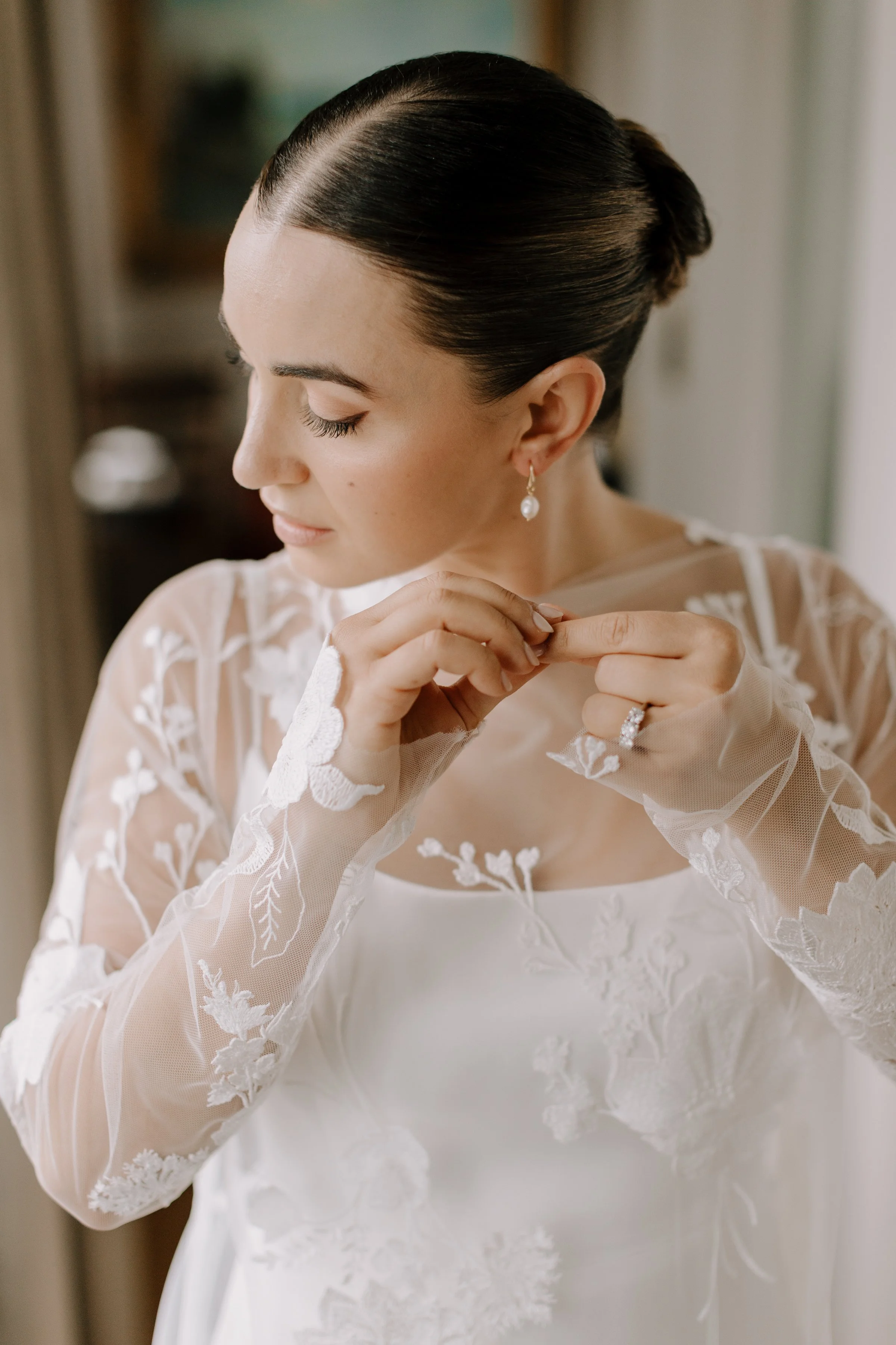 A woman in a white wedding dress with lace details, wearing pearl earrings, adjusting her dress with her eyes closed, indoors with soft lighting.