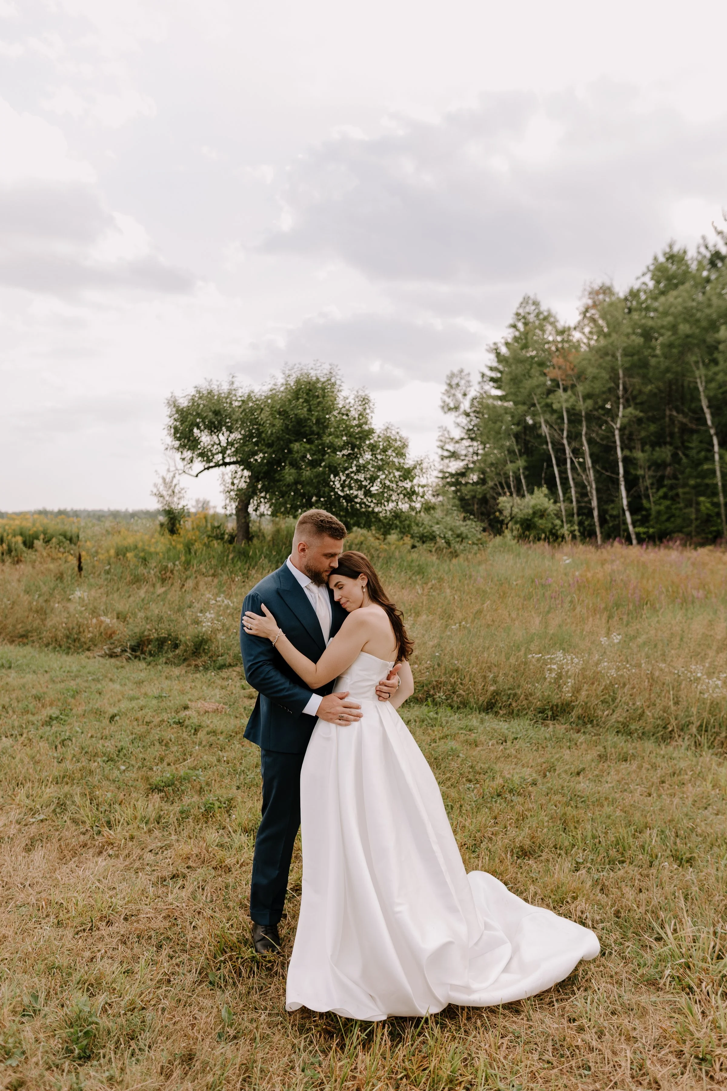 A bride and groom embracing outdoors in a grassy field with trees and overcast sky in the background.