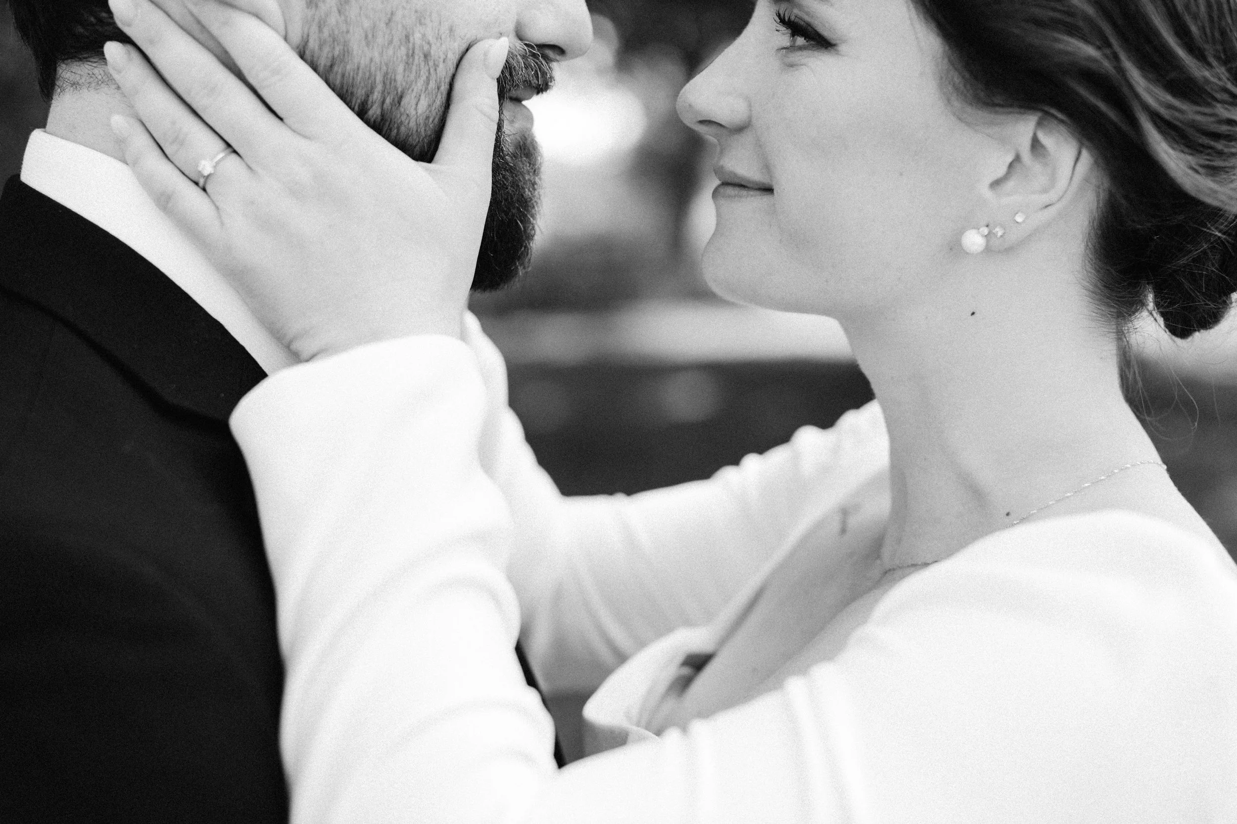 A black-and-white photograph of a couple close together. The woman gently touches the man's face, and they are gazing into each other's eyes in an intimate moment, possibly during their wedding or a special occasion.
