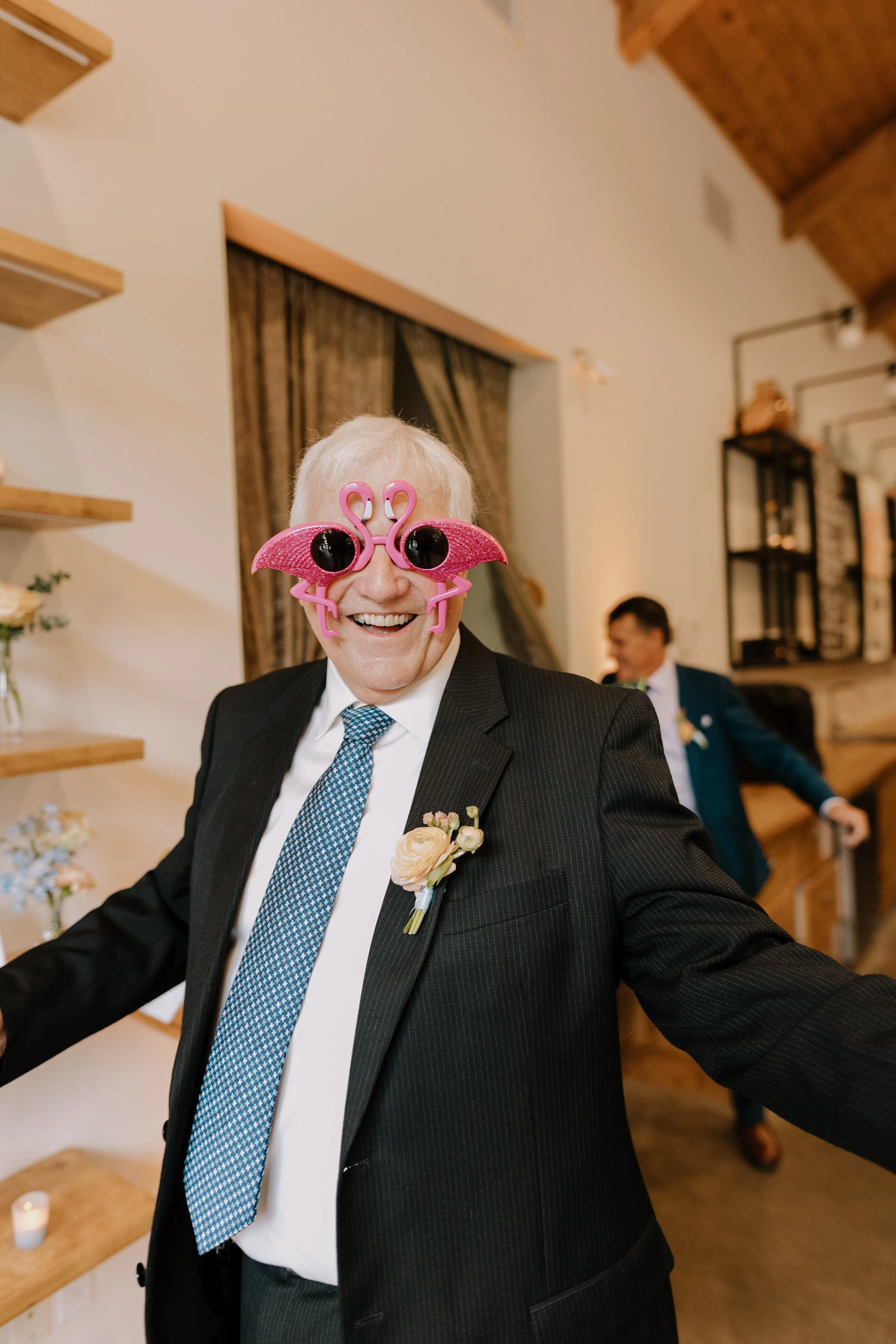 Elderly man in black suit and tie wearing pink flamingo sunglasses at a festive gathering.