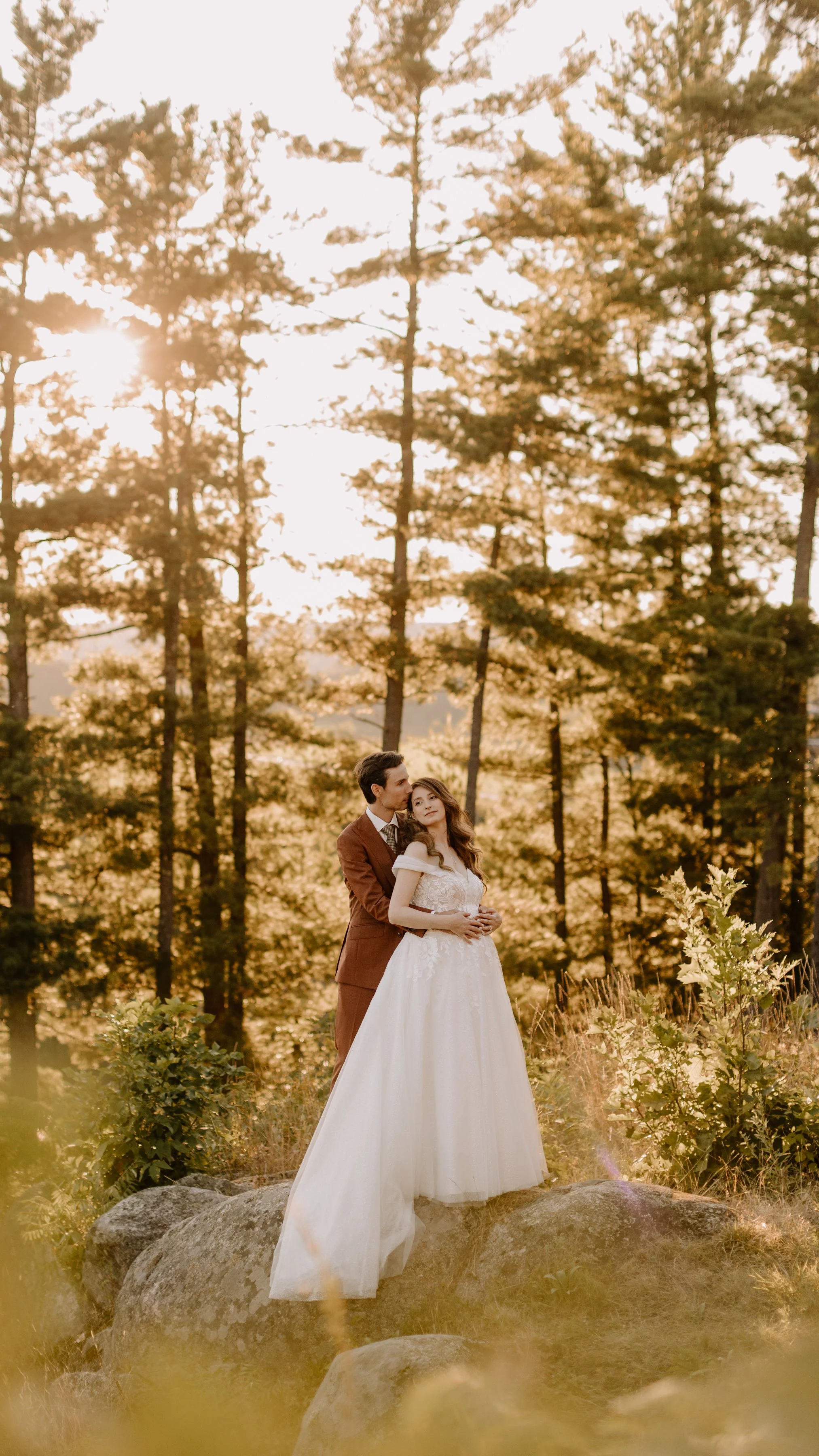 A bride in a white wedding dress and a groom in a brown suit standing on rocks in a forest with tall pine trees at sunset.