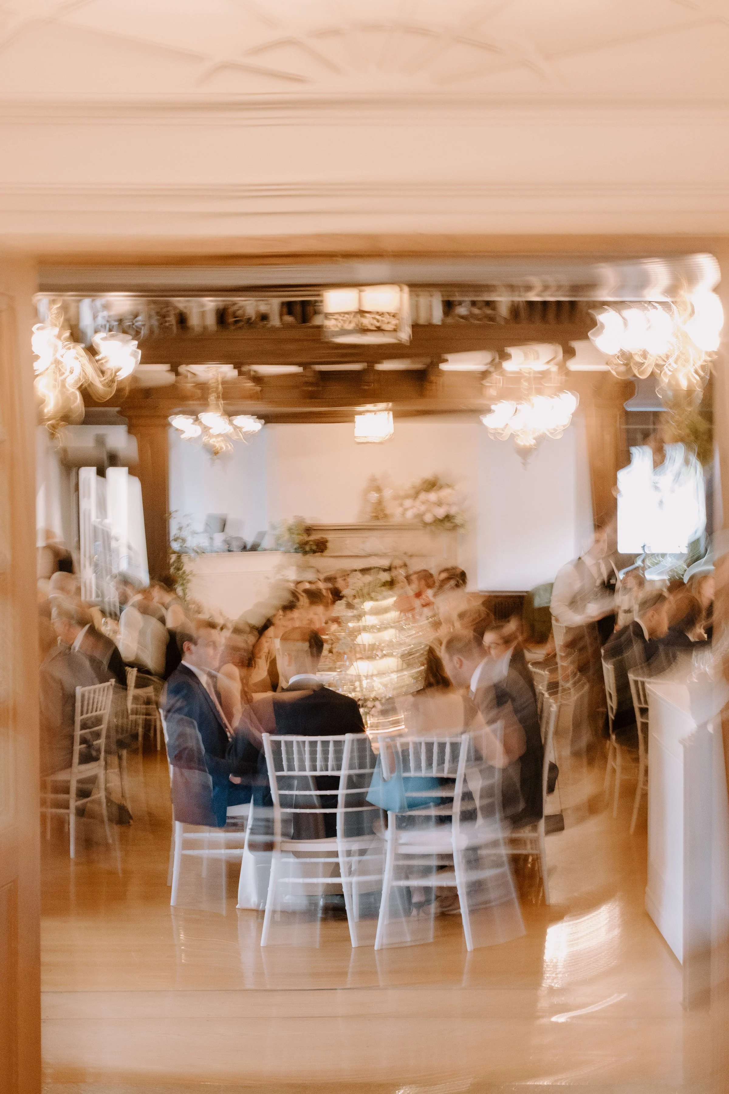 A banquet hall filled with people seated at round tables, dressed formally, with chandeliers hanging from the ceiling and a decorated stage in the background.