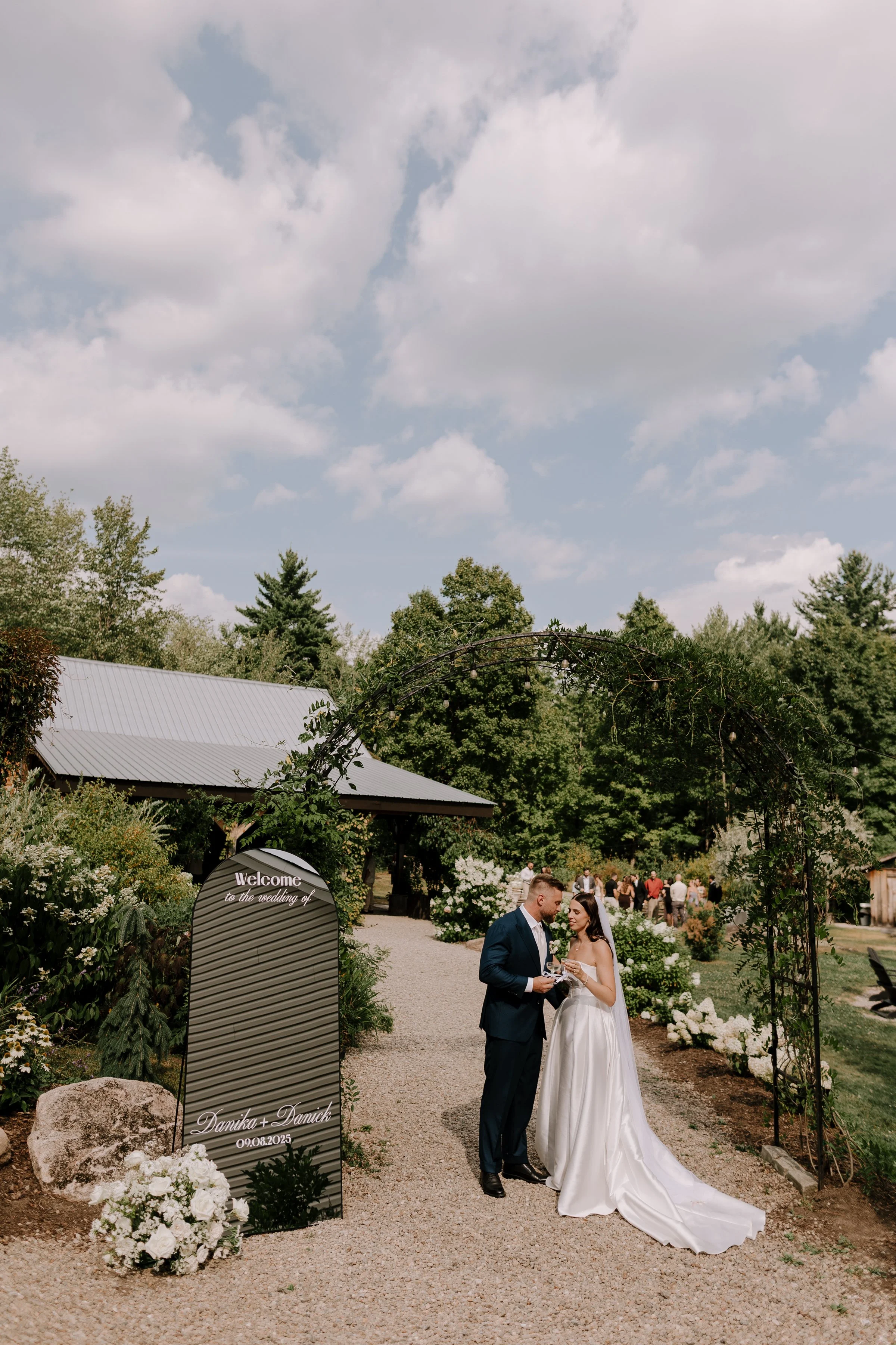A newlywed couple in wedding attire standing hand in hand outdoors at a garden wedding, exchanging vows or having a moment together, surrounded by other guests and floral decorations.