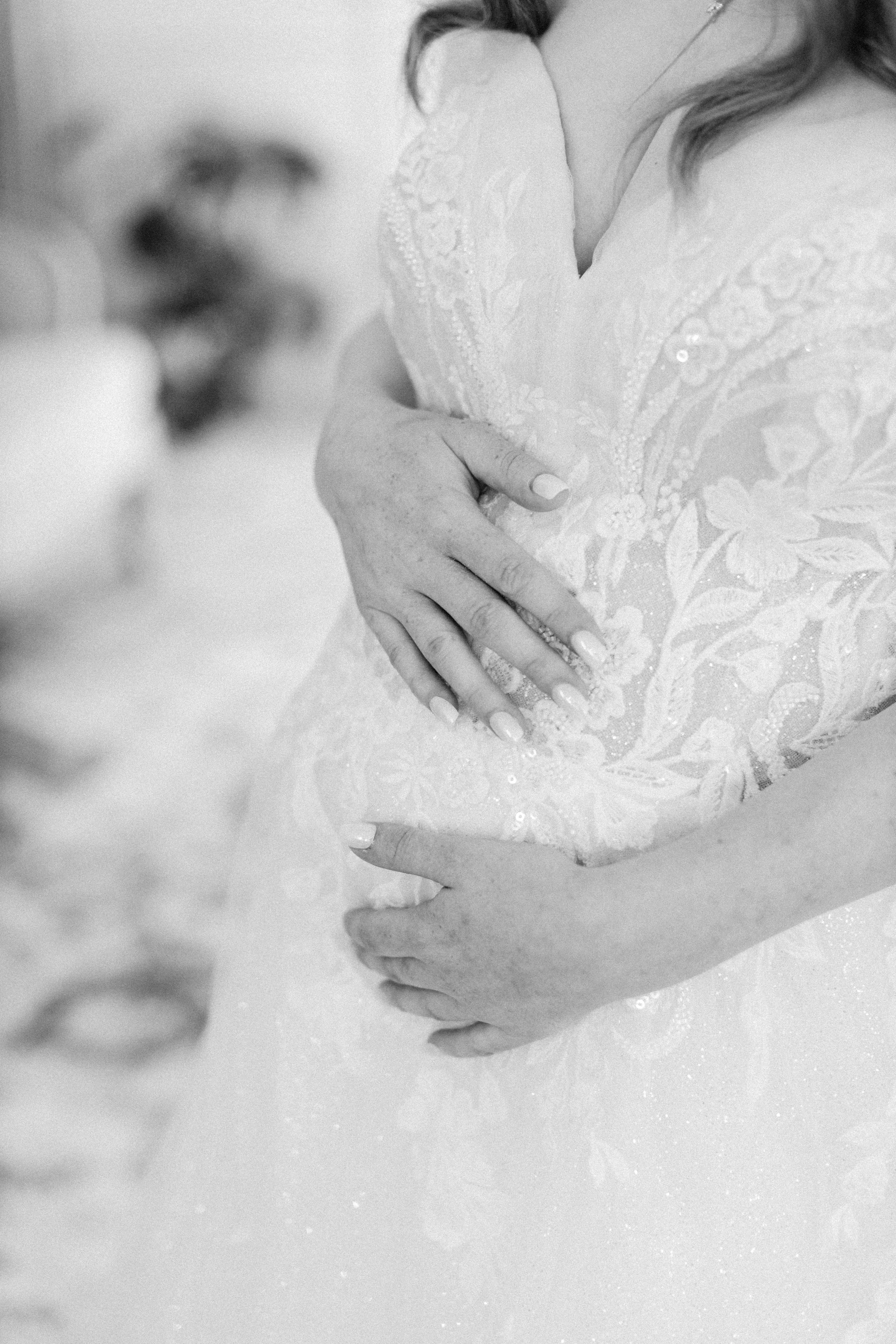 Close-up of two hands gently holding each other, one hand appears to be a bride's, showing well-manicured nails, and lace fabric of a wedding dress visible.