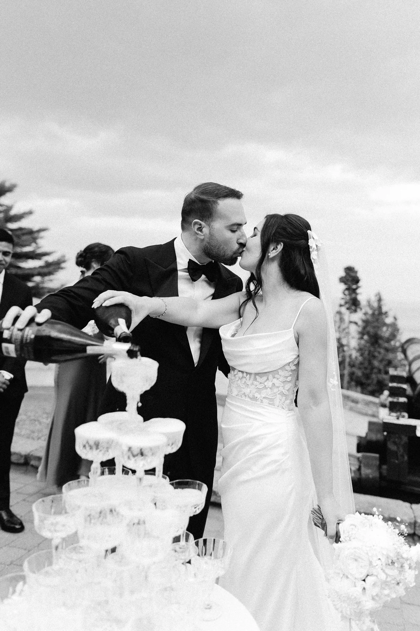 A black and white photo of a bride and groom kissing at their wedding reception, with a wedding cake in the foreground and a tiered cake stand visible. The bride is wearing a strapless lace dress, and the groom is in a tuxedo with a bow tie. There are wedding guests in the background, outdoors with trees and an overcast sky.