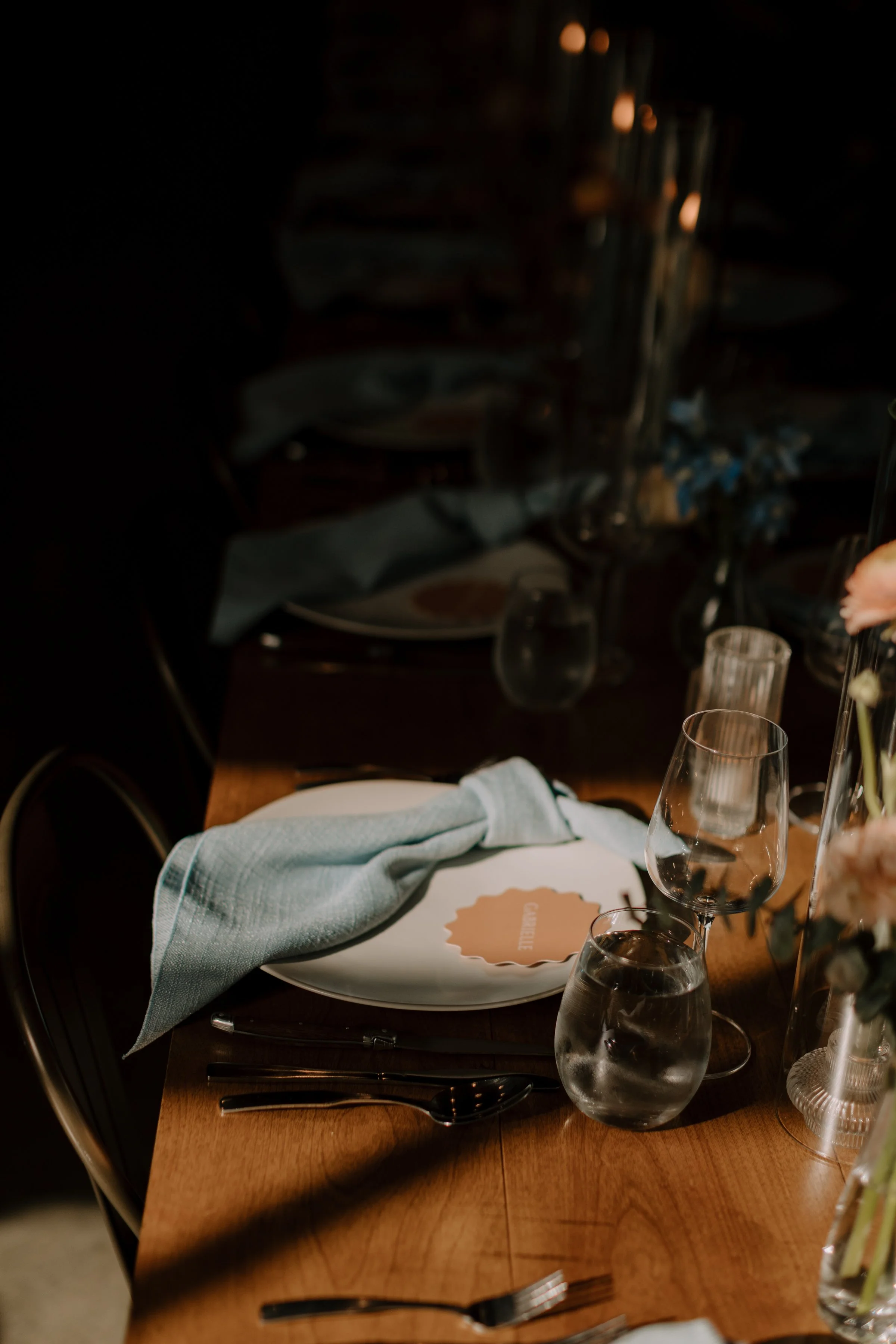 A dining table set with glasses of water, a white plate, a cloth napkin, and cutlery. In the background, there are decorative elements and dim lighting.