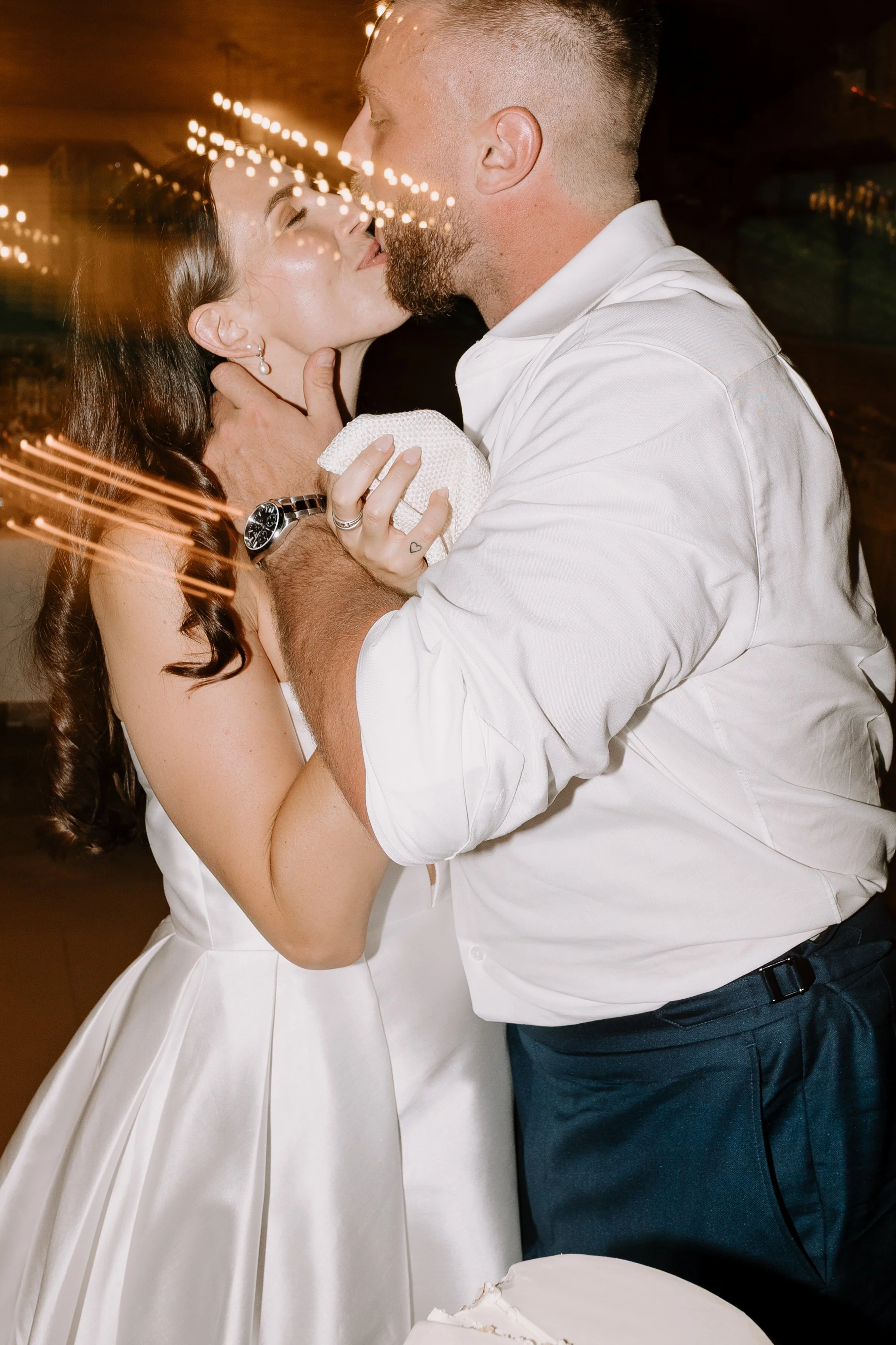 A couple kissing at a wedding reception, with the bride wearing a white strapless dress and the groom in a white shirt and dark pants. The bride has long brown hair and is holding a small white cake. The groom has short hair and a beard. The backgrou
