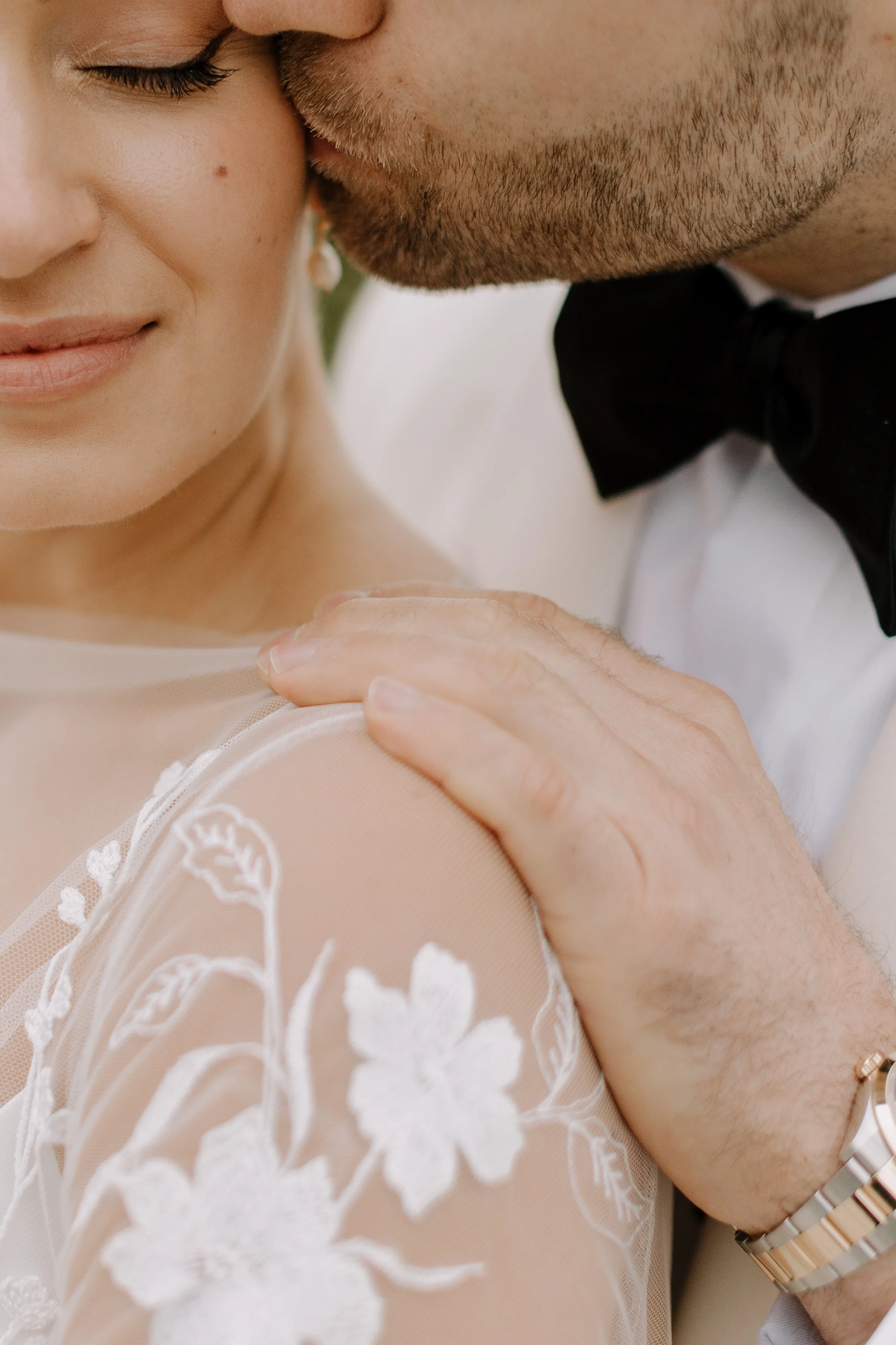 A close-up of a bride and groom sharing a kiss, with the groom gently holding the bride's shoulder. The bride is wearing a white lace dress with floral embroidery, and the groom is dressed in a tuxedo with a black bow tie and a gold bracelet on his w