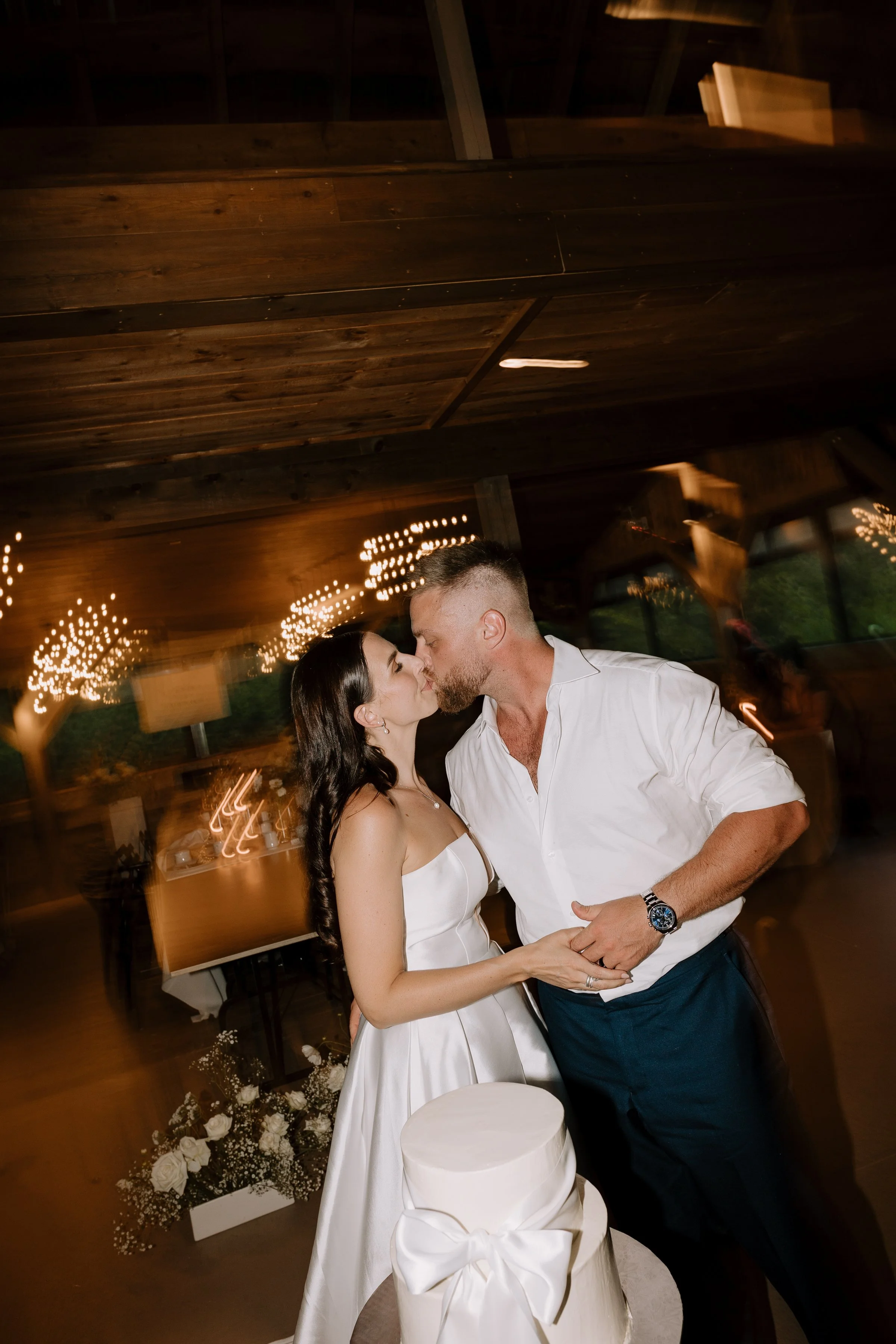 A couple dancing closely at a wedding reception, with romantic expressions and holding hands, in a decorated venue with string lights and floral arrangements.
