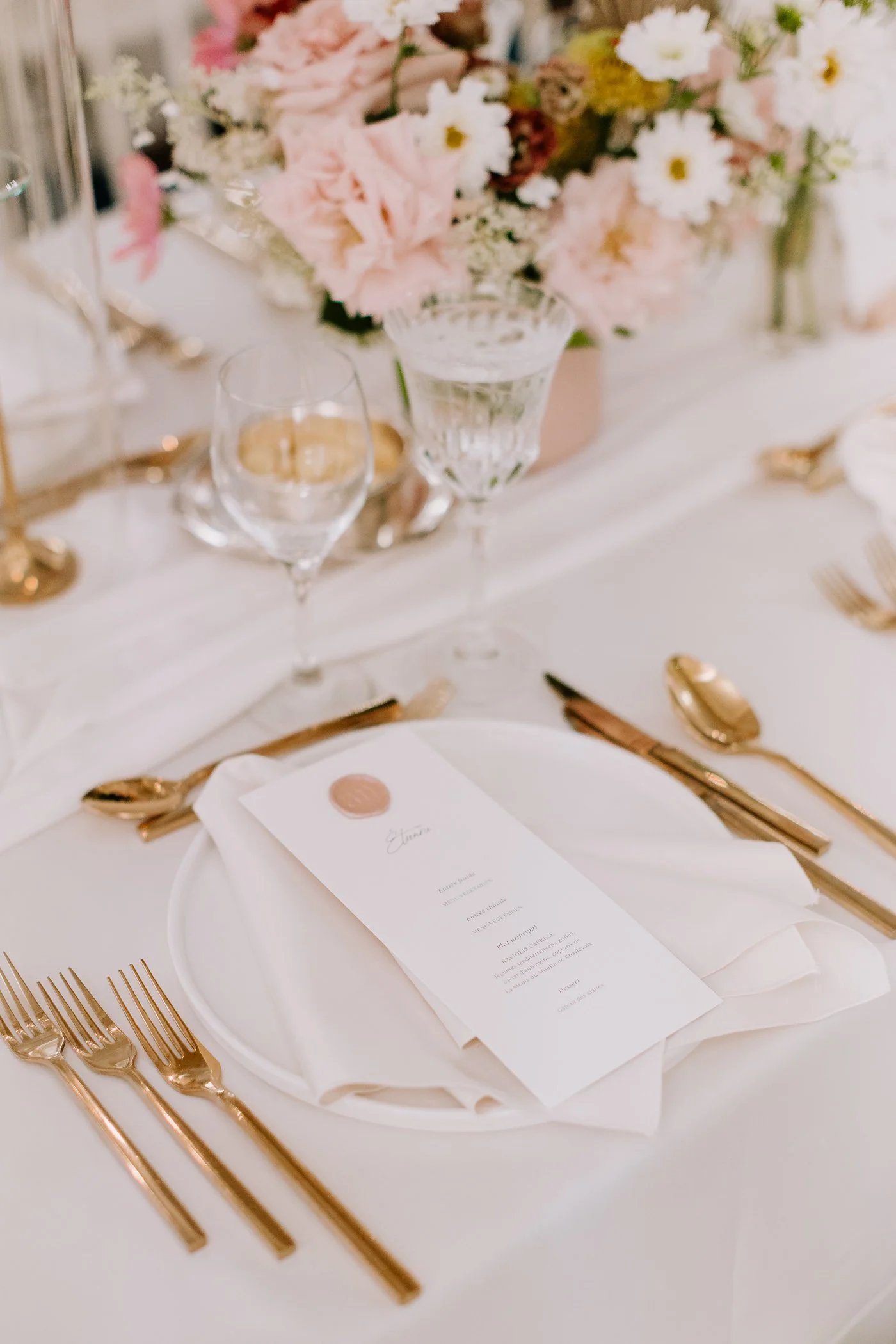 A formal table setting with gold utensils, a white plate with a folded napkin, and a printed menu on top. In the background, there is a floral centerpiece with pink and white flowers, and two glasses of water.
