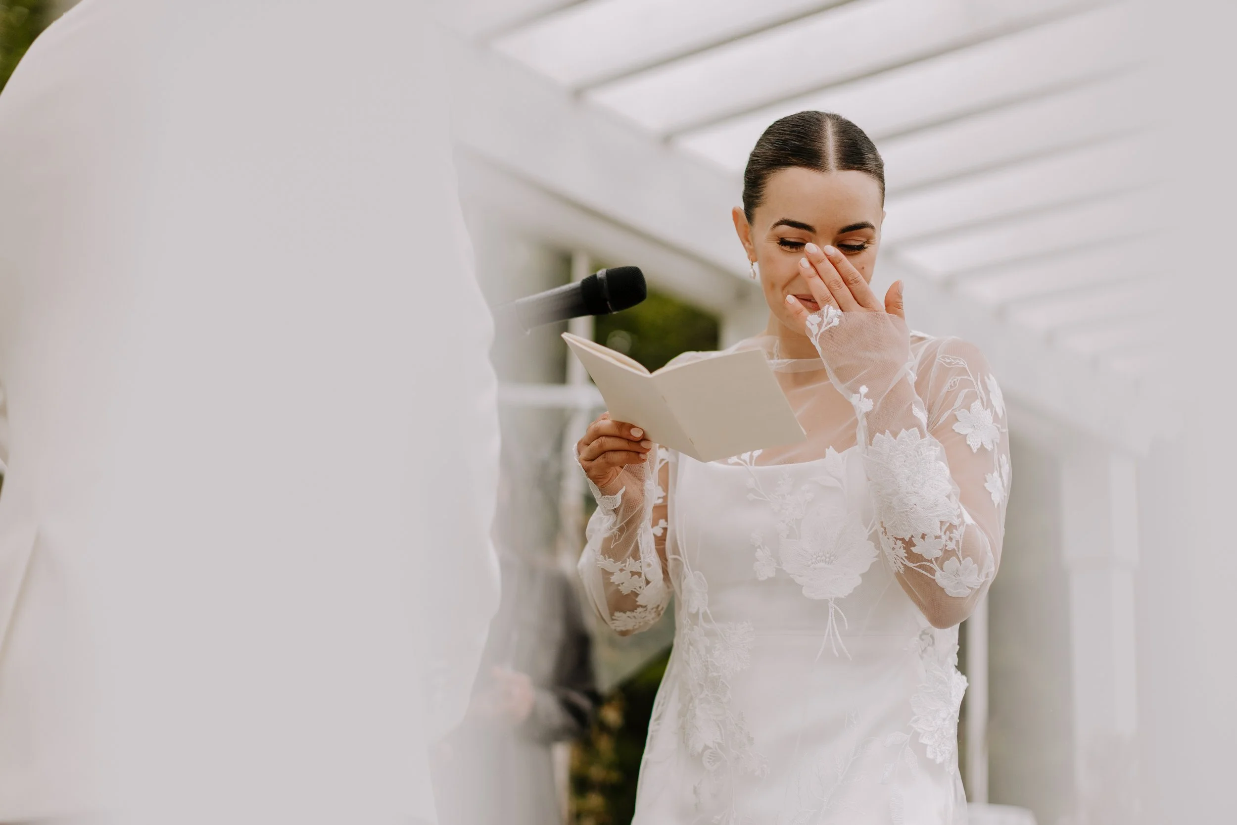 A woman in a white lace wedding dress wiping tears with her hand, standing outdoors under a white pergola.