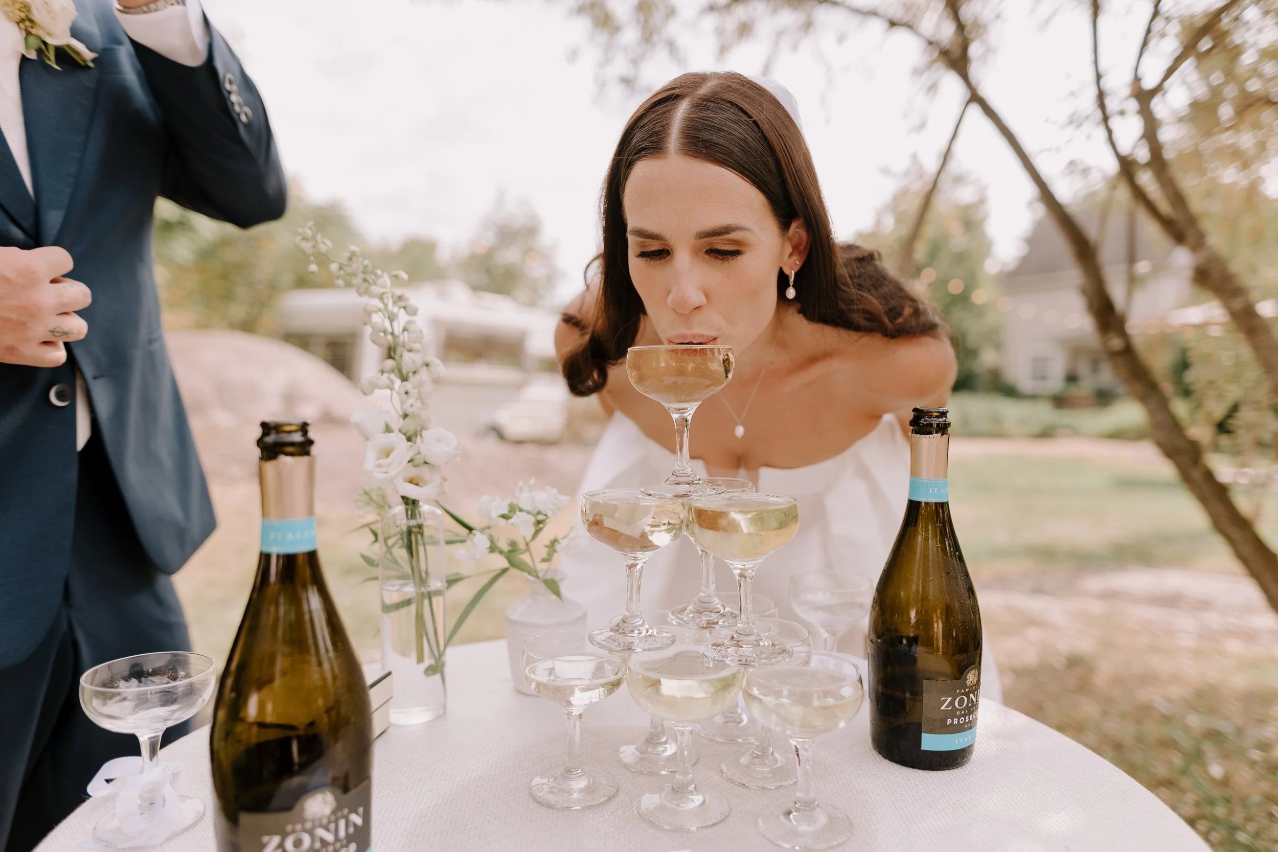 Woman in wedding dress drinking champagne from a glass under a pyramid of champagne glasses at an outdoor wedding reception.