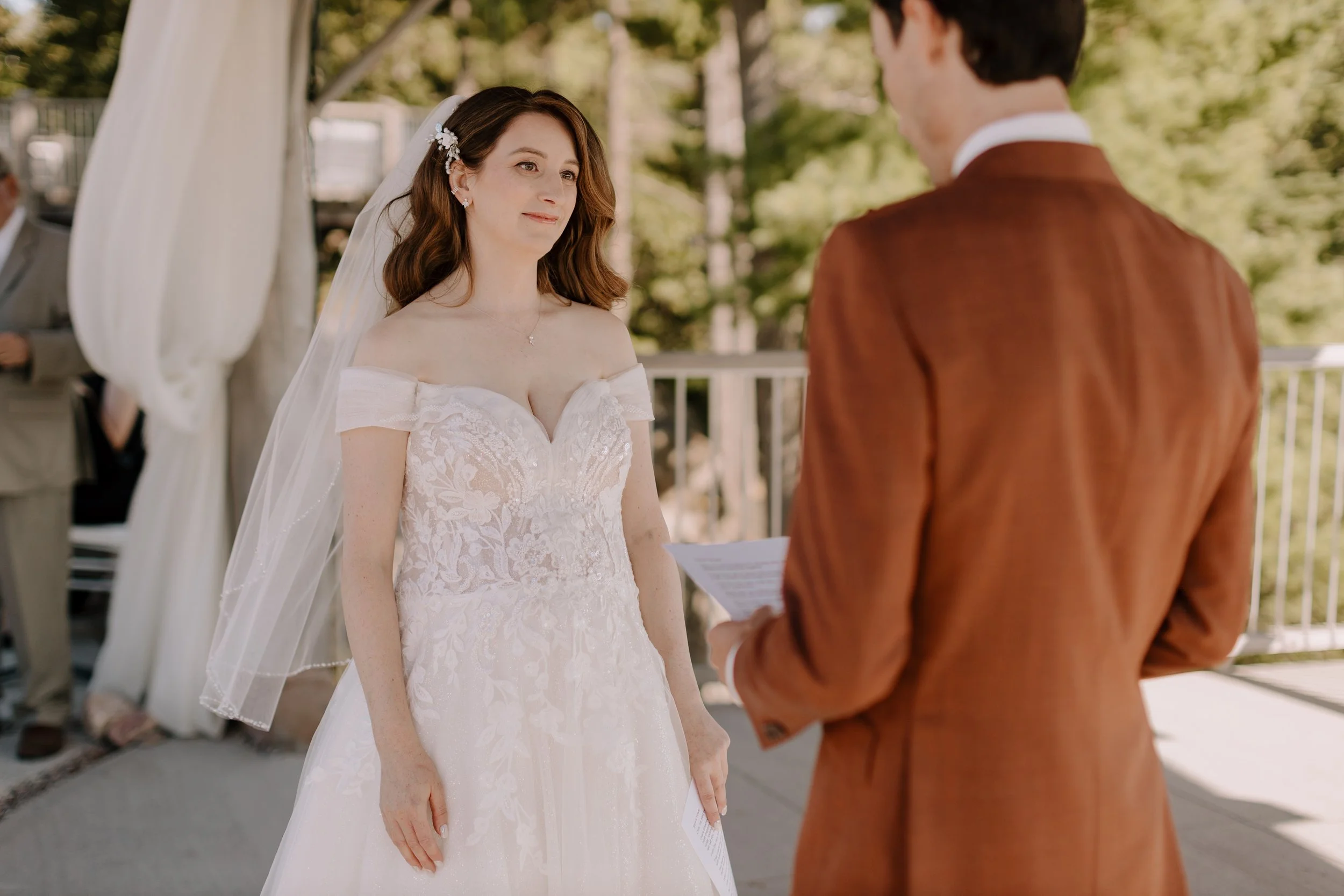 A bride with long brown hair in a white lace wedding dress and a veil stands facing a groom during a wedding ceremony outdoors, with a man in a brown suit holding a paper in front of a backdrop of trees and a white tent.