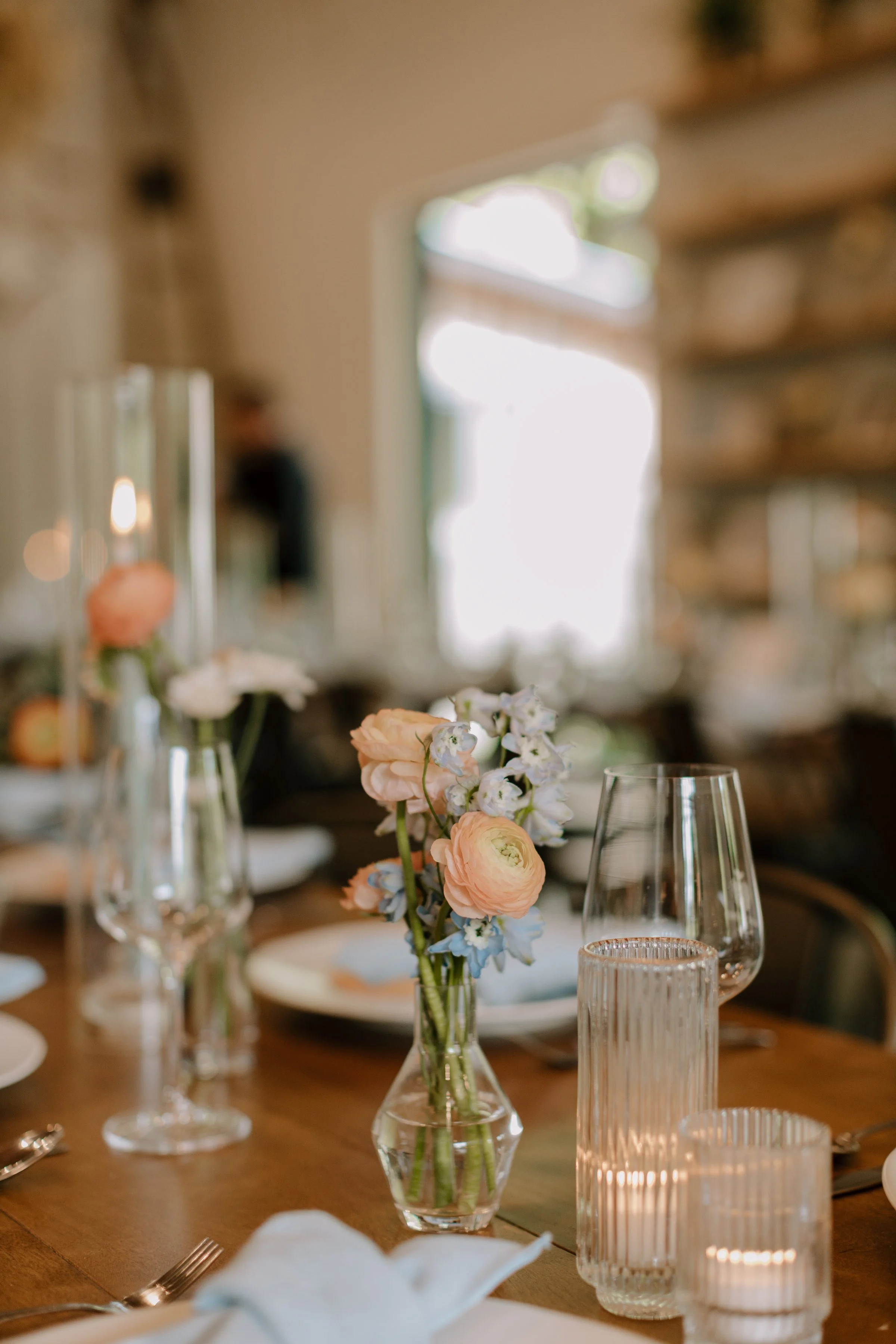 A dining table decorated with vases of pink and blue flowers, surrounded by glassware including wine glasses, and candles in ribbed holders. The room has exposed brick walls and a large window letting in natural light.