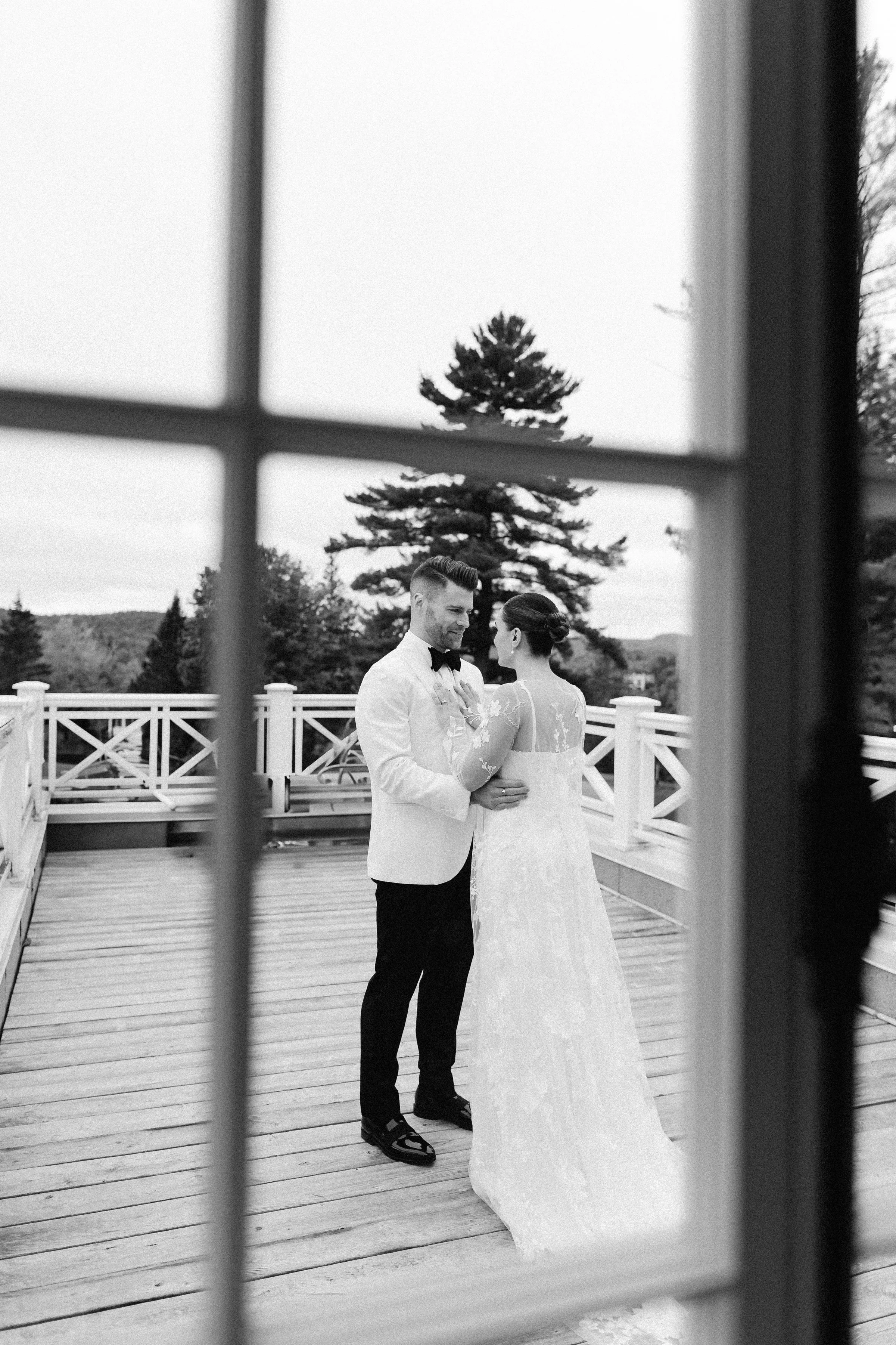 A black and white photo of a couple dressed in wedding attire, standing on a wooden deck outdoors. The scene is viewed through window or door frames, with trees and a cloudy sky in the background.