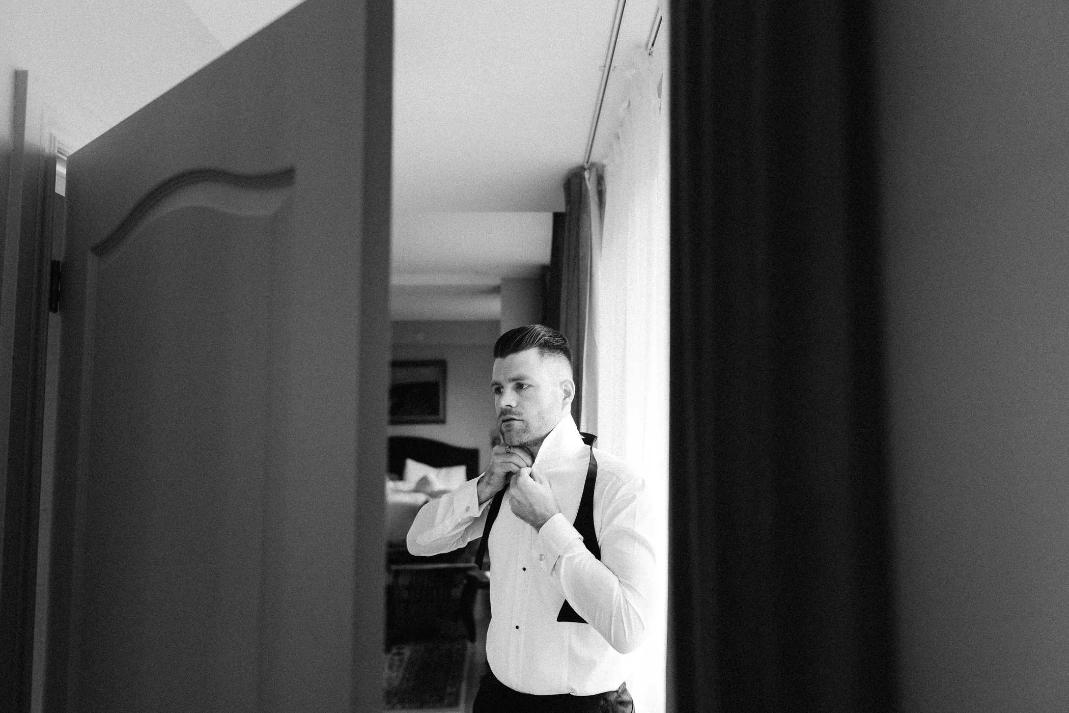 A man dressed in formal attire, adjusting his collar in front of a mirror, with a window and curtains in the background.
