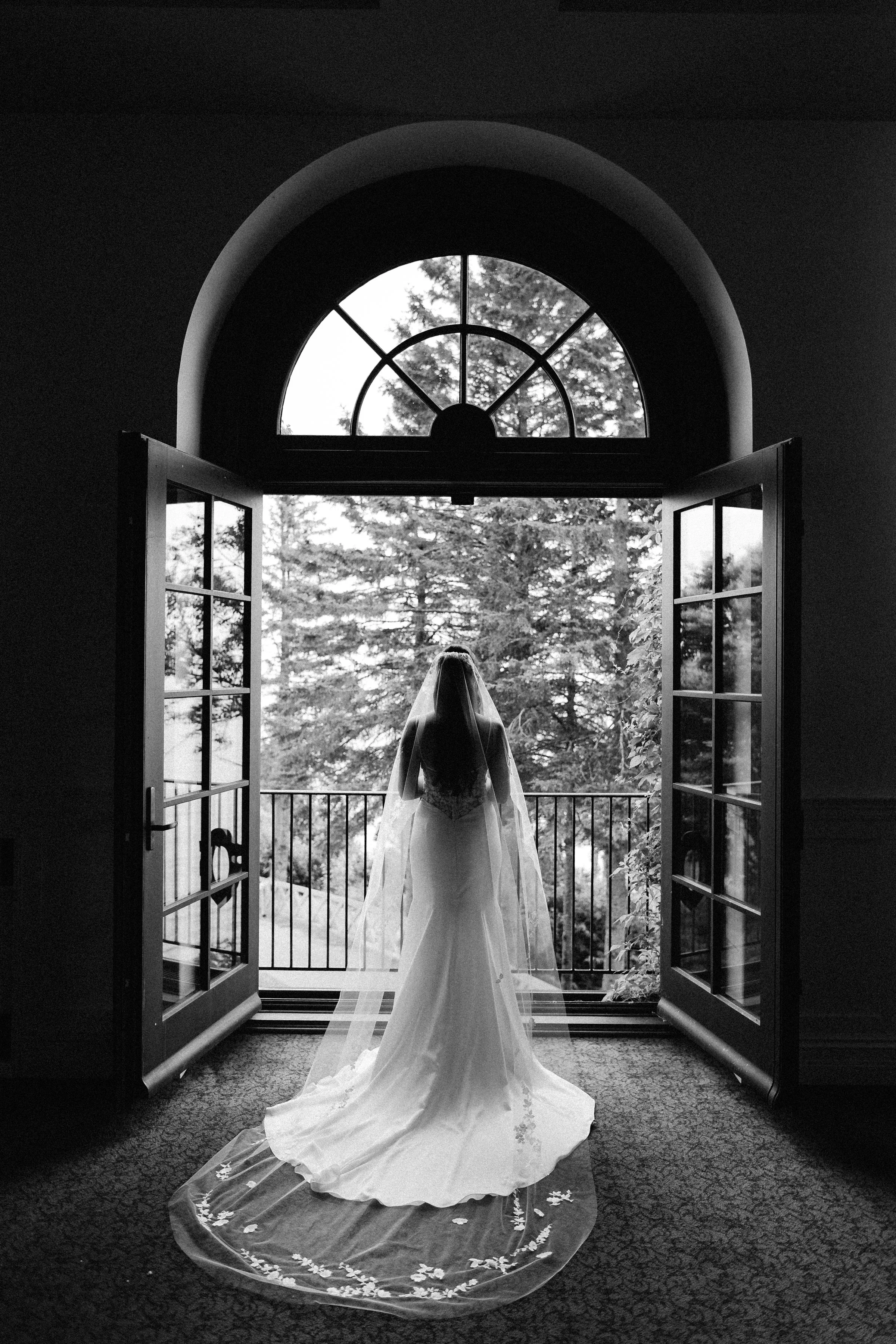 A bride standing at an open door with a veil and wedding gown, looking out to a forested area outside.
