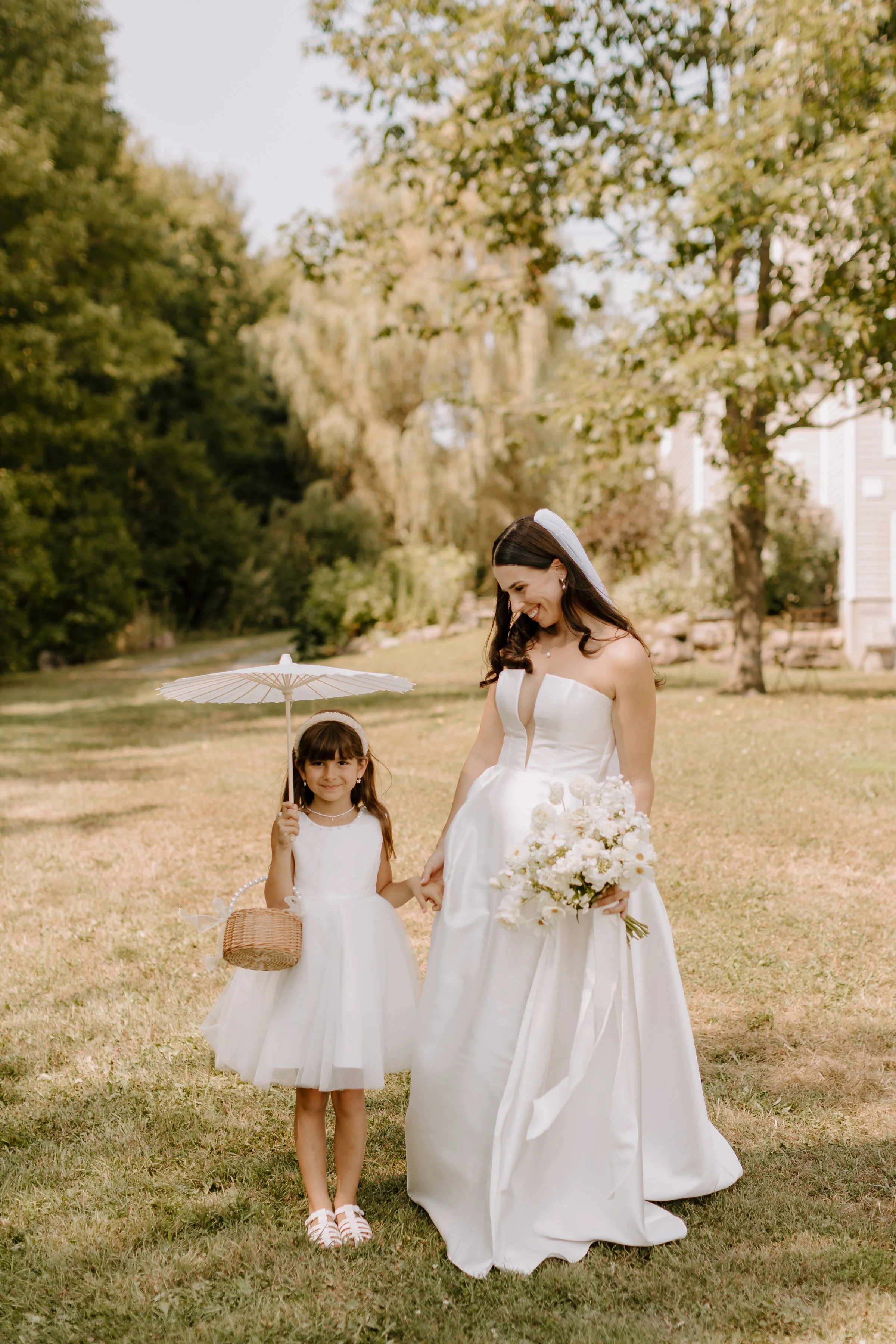 A woman in a wedding dress holding a bouquet and a young girl in a white dress carrying a basket and holding a parasol, walking in a park with green trees and grass.