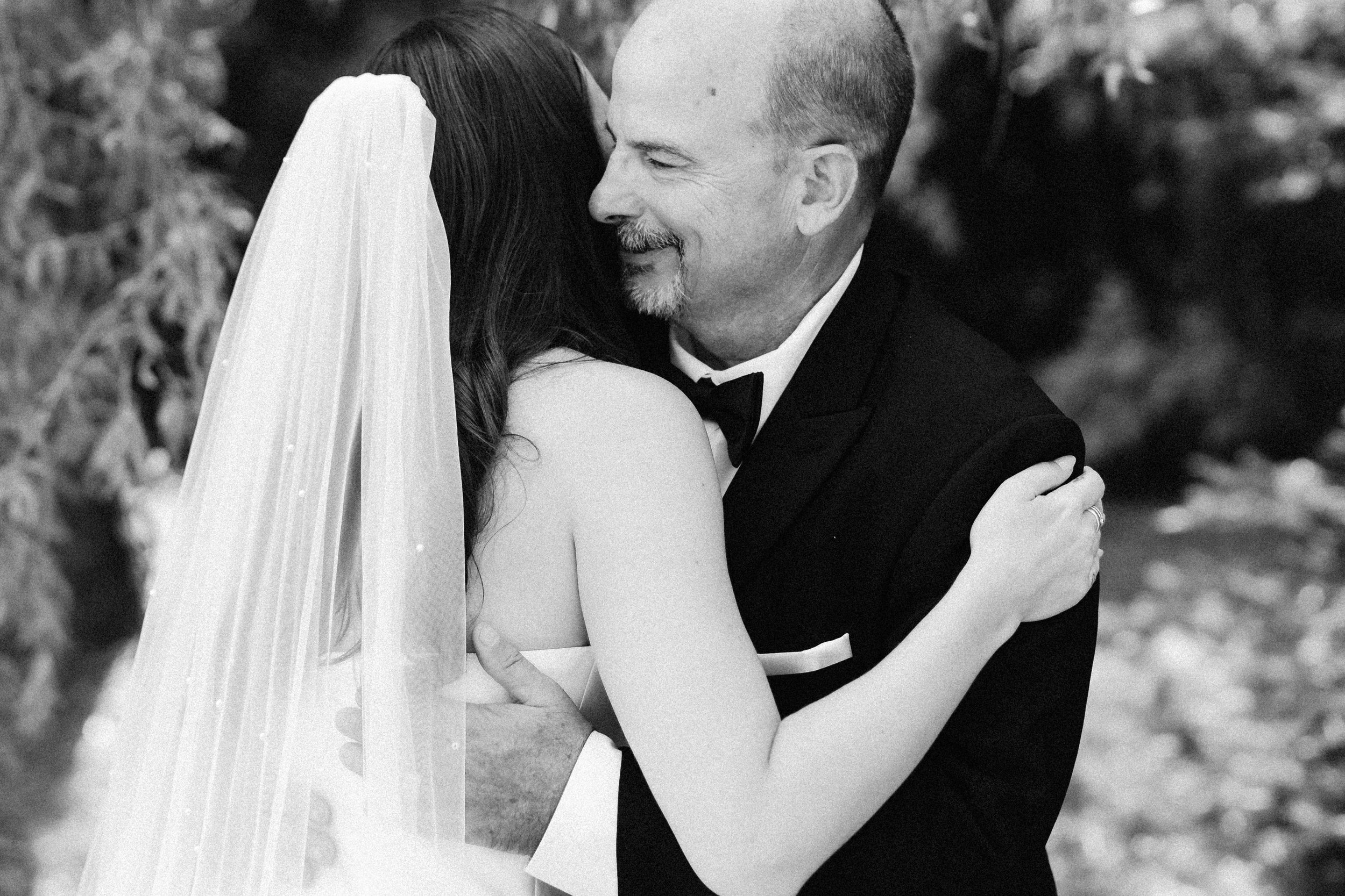 A black-and-white photograph of a couple kissing, with the woman wearing a veil and the man in a tuxedo. The woman has long dark hair, and the man has short hair and a beard. The background appears to be outdoors with blurred foliage.