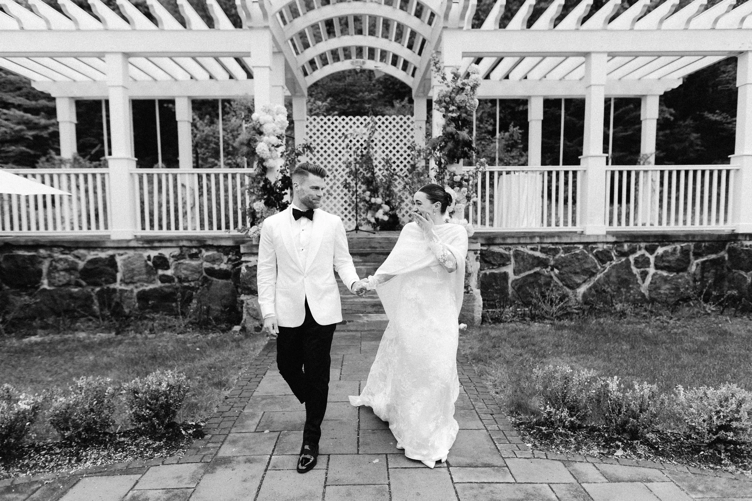 A black and white photo of a bride and groom holding hands outdoors. The groom is wearing a white tuxedo jacket with a black bow tie, and the bride is in a long white wedding dress with lace sleeves. They are walking on a stone path in front of a gaz