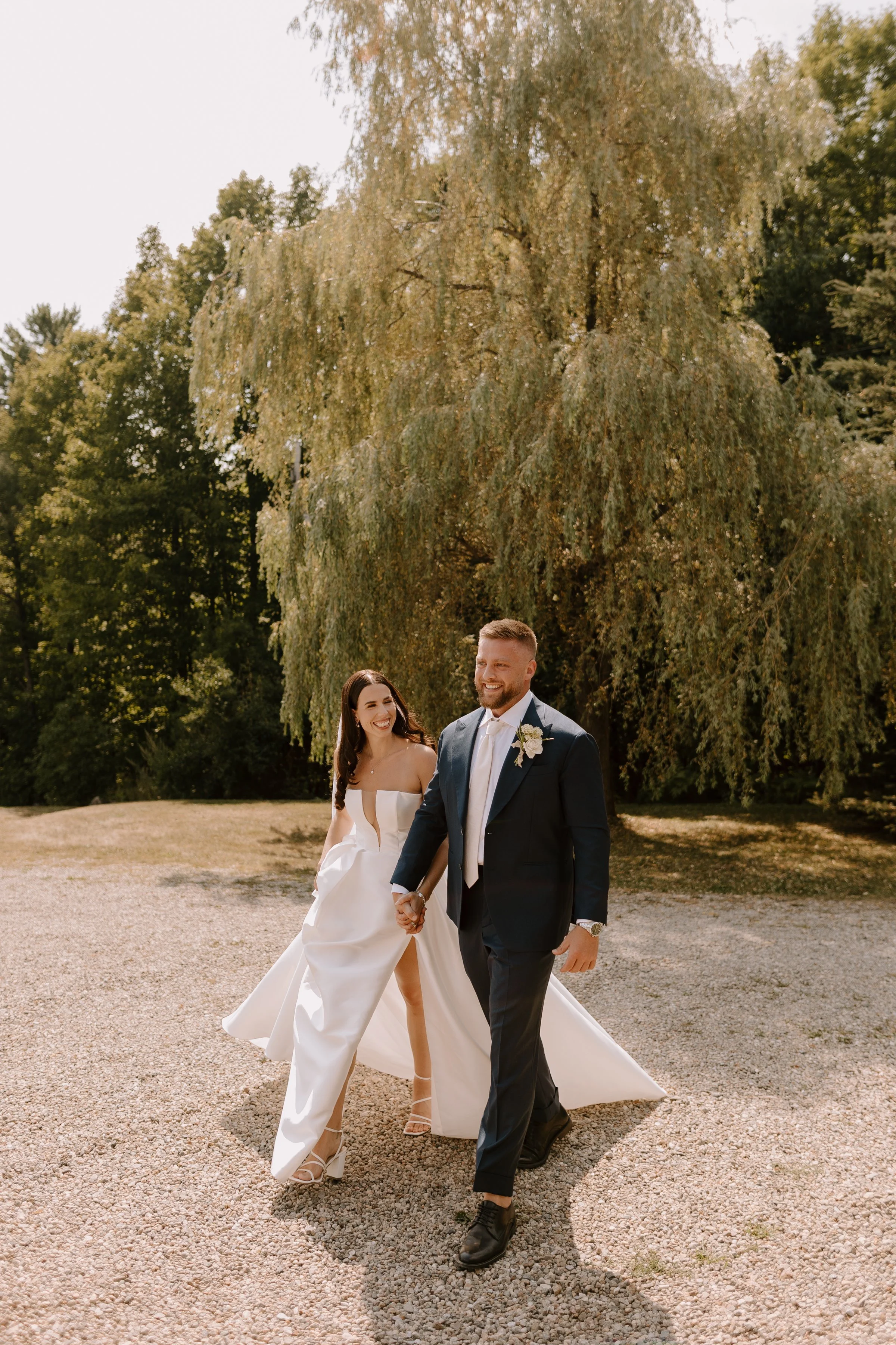 A newlywed couple holding hands and walking outdoors beneath a large leafy tree, with the bride smiling and wearing a strapless wedding dress and heels, and the groom smiling and dressed in a dark suit and tie.