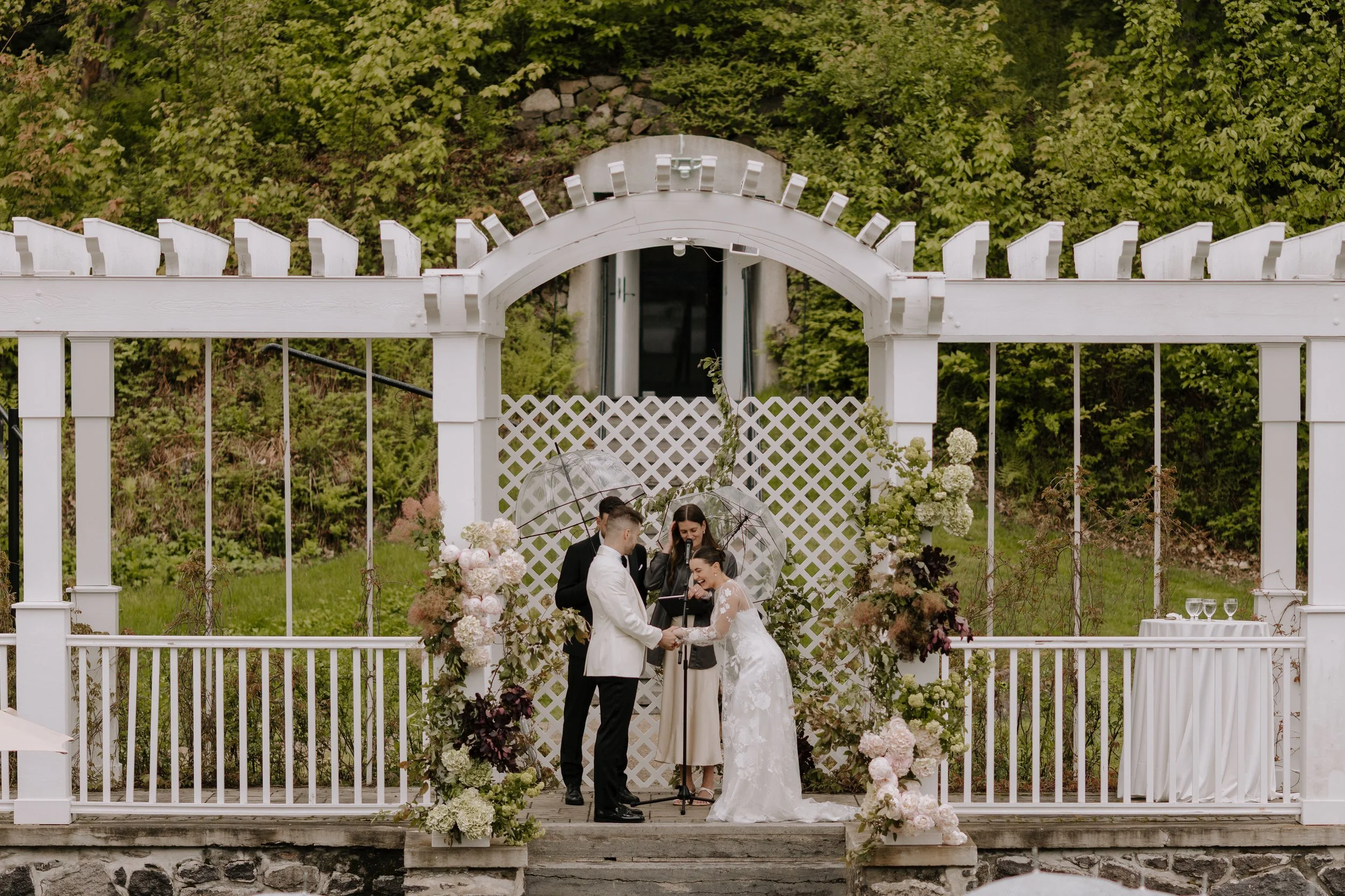 A wedding ceremony taking place outdoors under a white pergola decorated with flowers, with a couple exchanging vows in front of an officiant and a small group of witnesses.