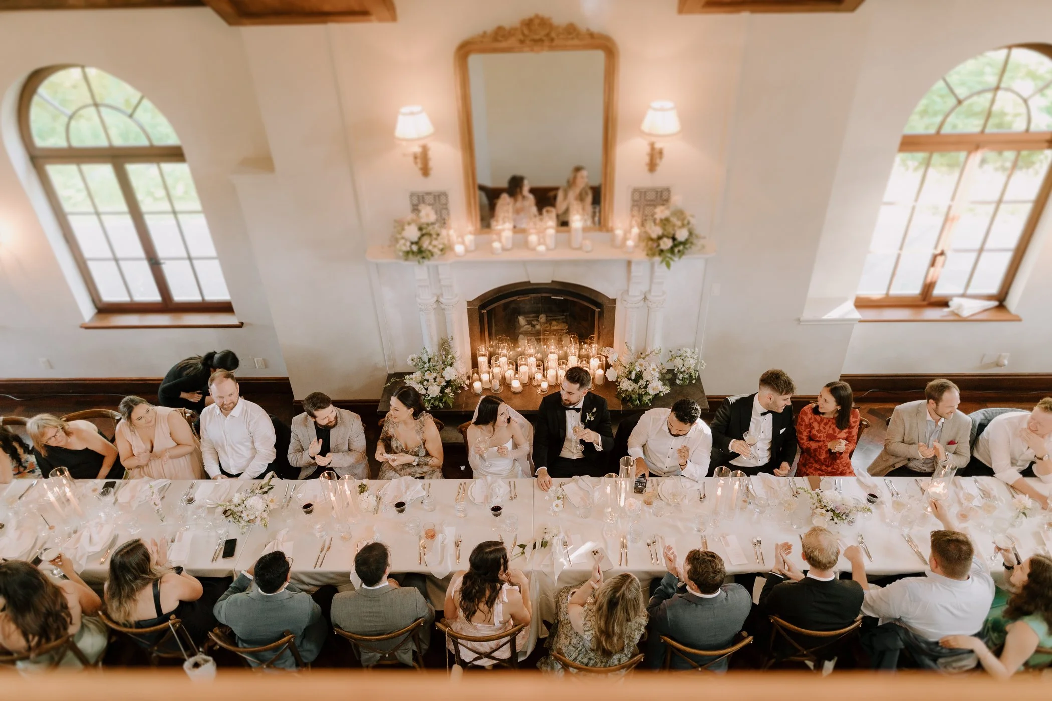 A banquet hall decorated for a wedding reception with a long head table where guests are seated, a fireplace with white flowers and candles, and large arched windows letting in natural light.