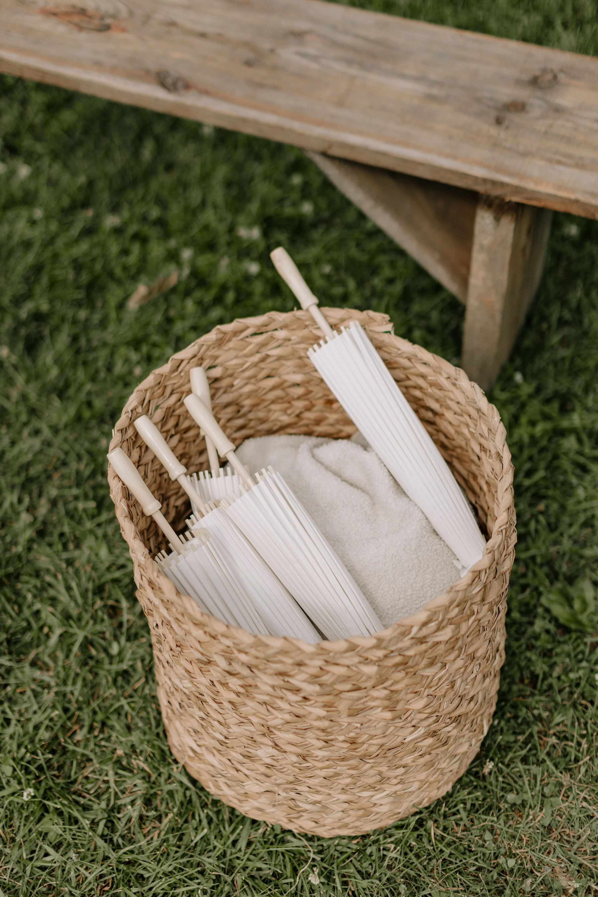 A woven basket containing white paper umbrellas and a white towel, placed on grass with a wooden bench in the background.