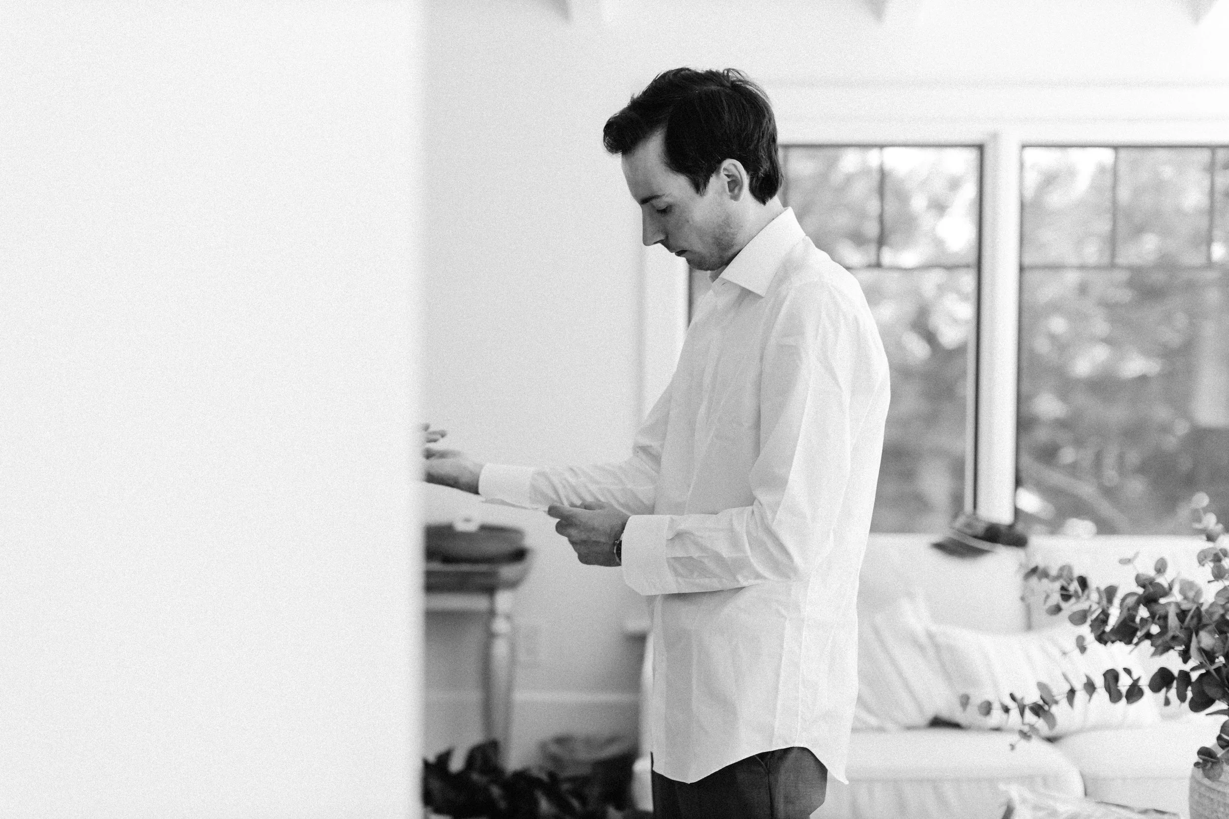 A man with dark hair and light skin wearing a white shirt, standing indoors near large windows with a view of trees outside. He is looking down at a paper he is holding in his hands.