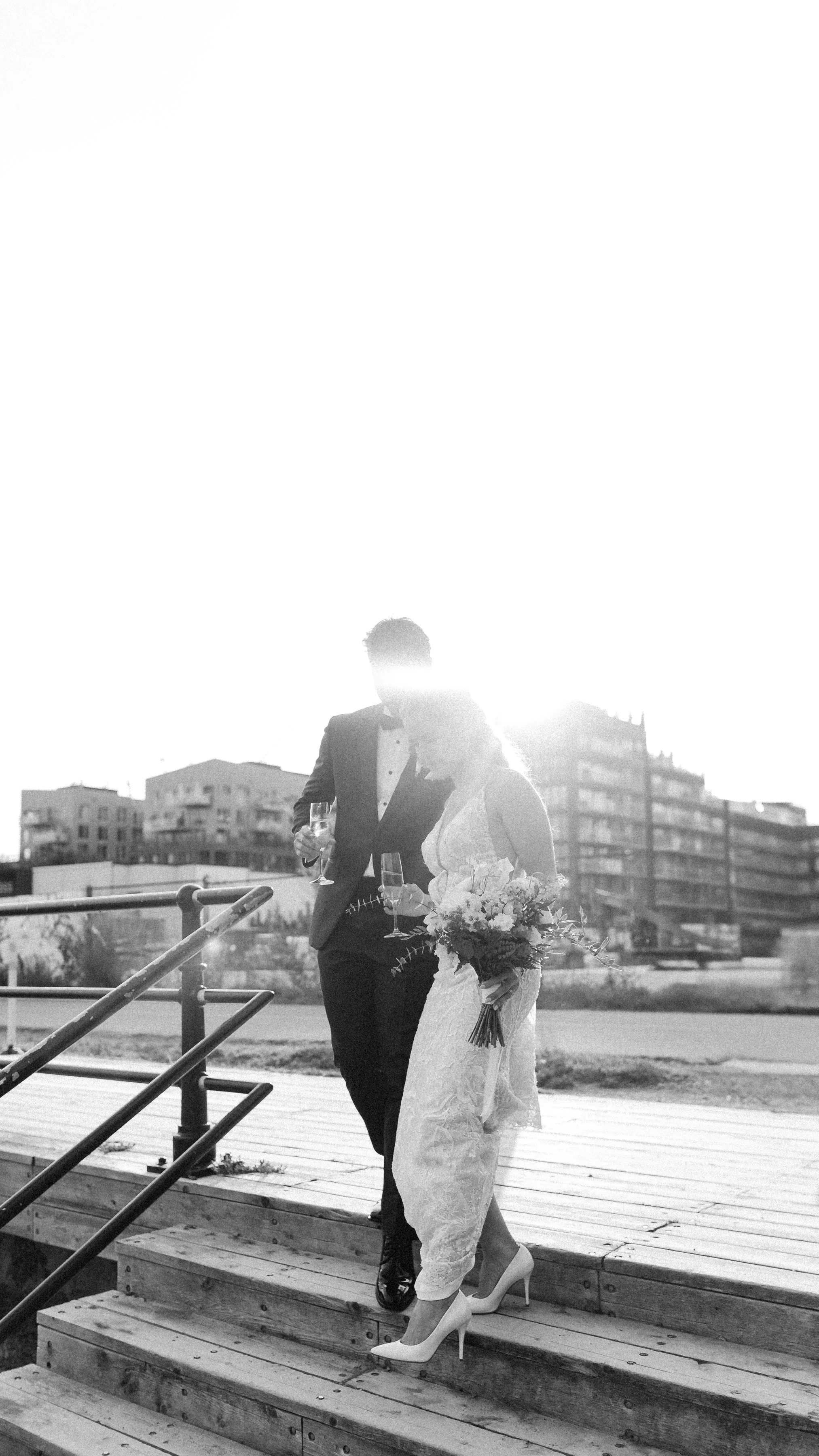 A black and white photo of a bride and groom walking down stairs at their wedding ceremony, holding glasses of champagne, with the sun behind them and city buildings in the background.