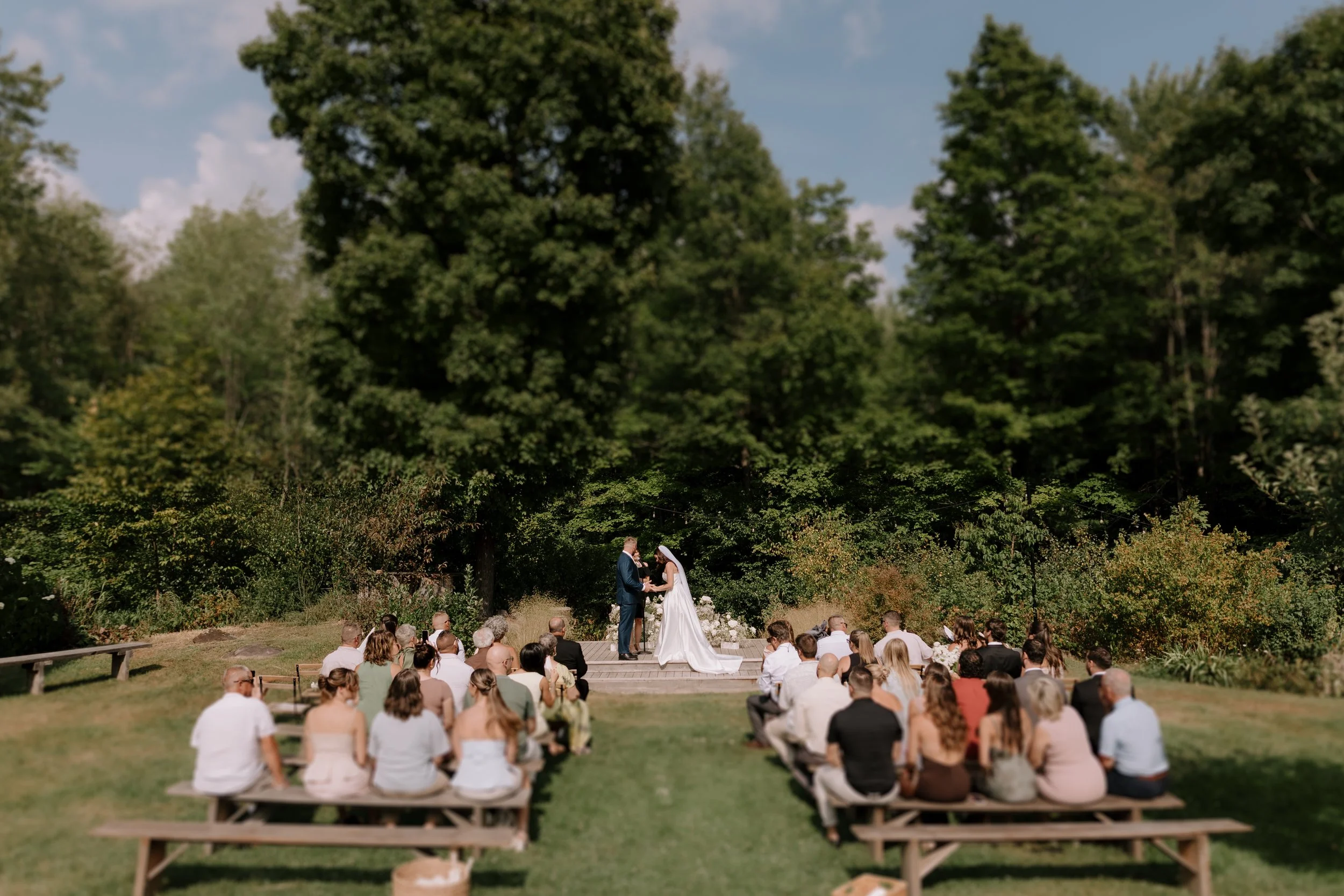 An outdoor wedding ceremony with a large audience seated on benches, a couple standing under an arch exchanging vows, surrounded by lush green trees and clear blue sky.