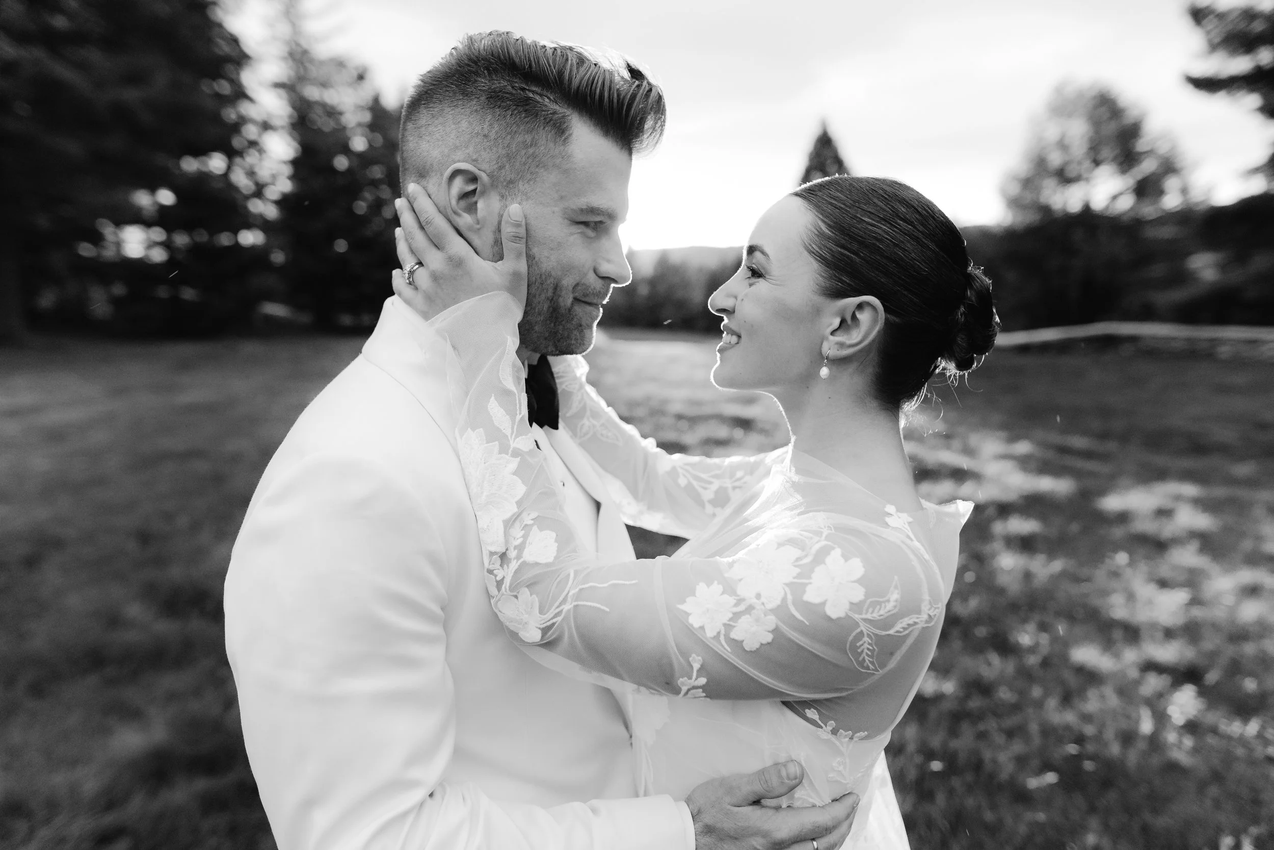 A black and white photo of a couple embracing outdoors, facing each other, with the man holding the woman around her waist. They are smiling and looking into each other's eyes, surrounded by trees and a partly cloudy sky.