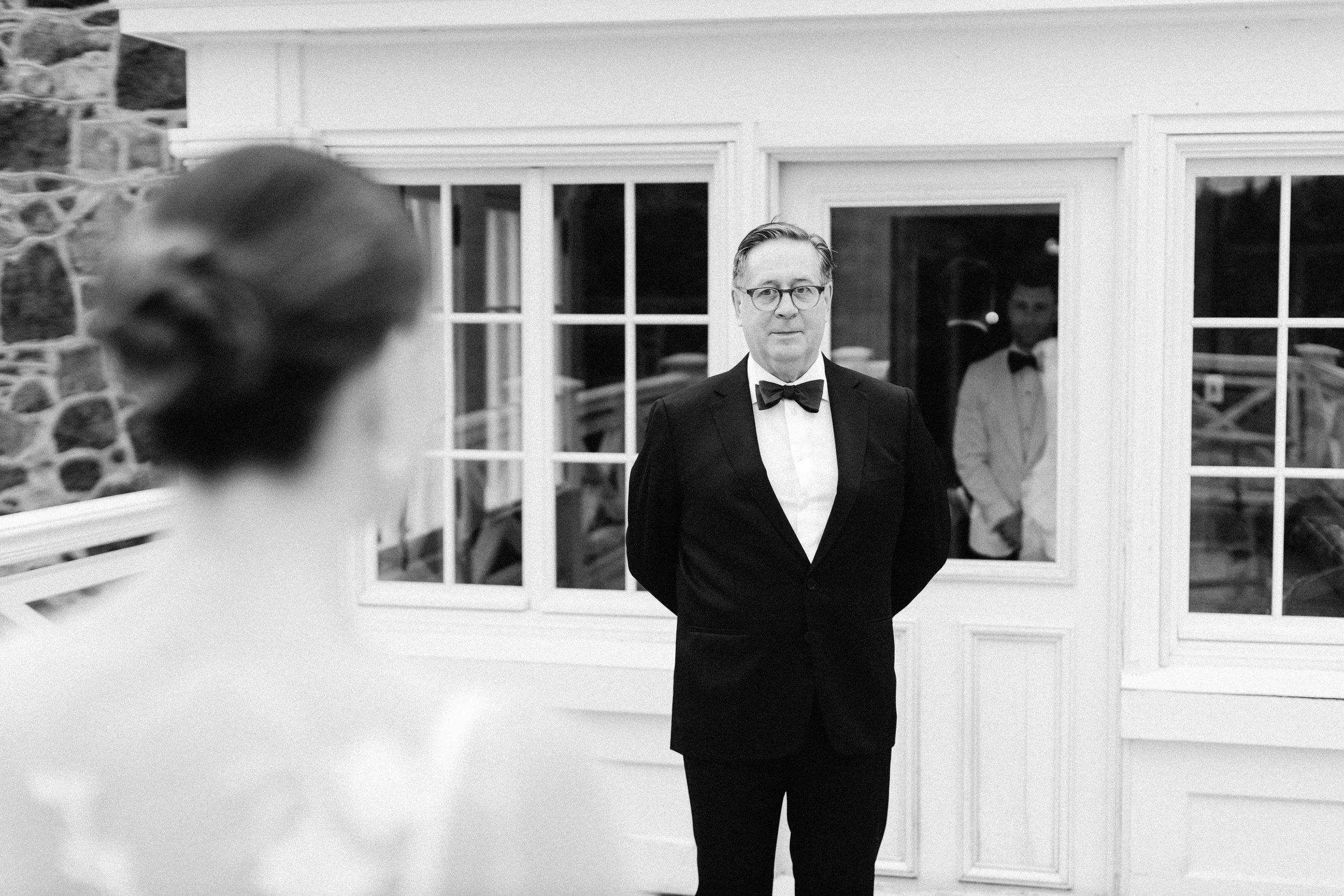 A man in a tuxedo with glasses standing indoors in front of a framed window with reflections of other people, and a woman in the background.