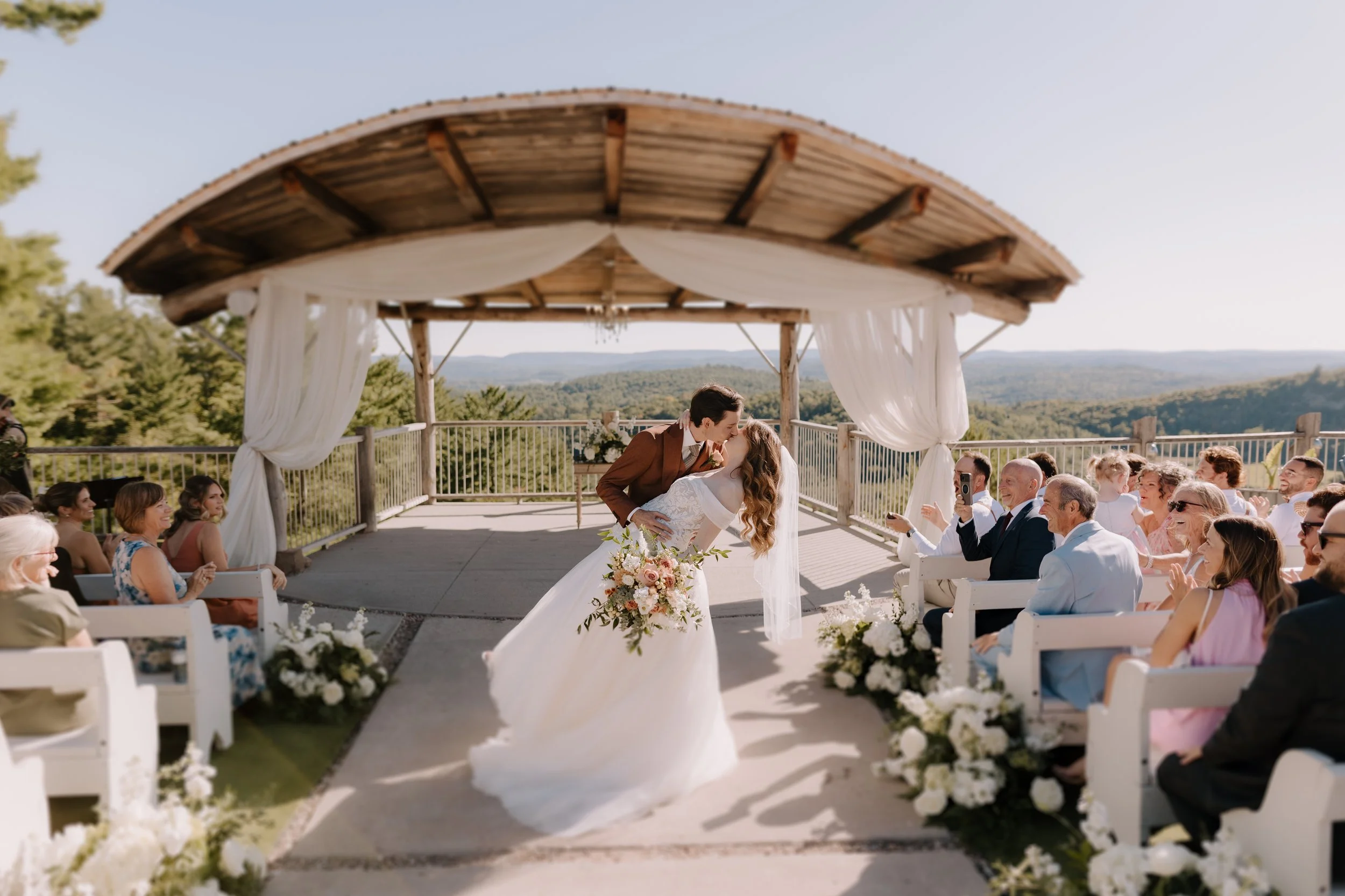 A bride and groom sharing a kiss during their outdoor wedding ceremony under a wooden arch with white drapery. Guests are seated on white benches, some smiling and taking photos, set against a scenic backdrop of green hills and a clear sky.