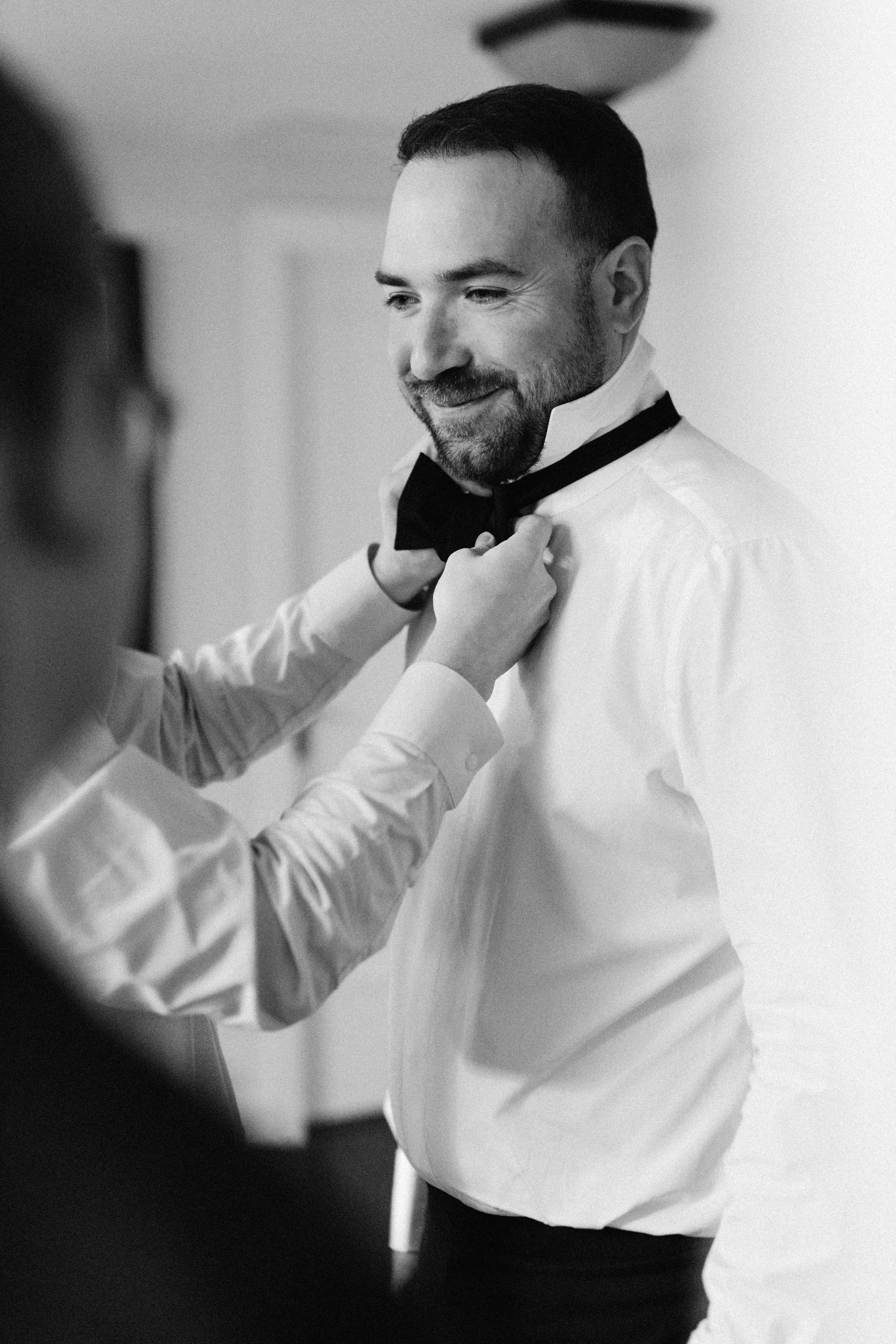 A man in formal attire is adjusting a bow tie on his shirt collar, smiling, as another person helps him in a well-lit room.