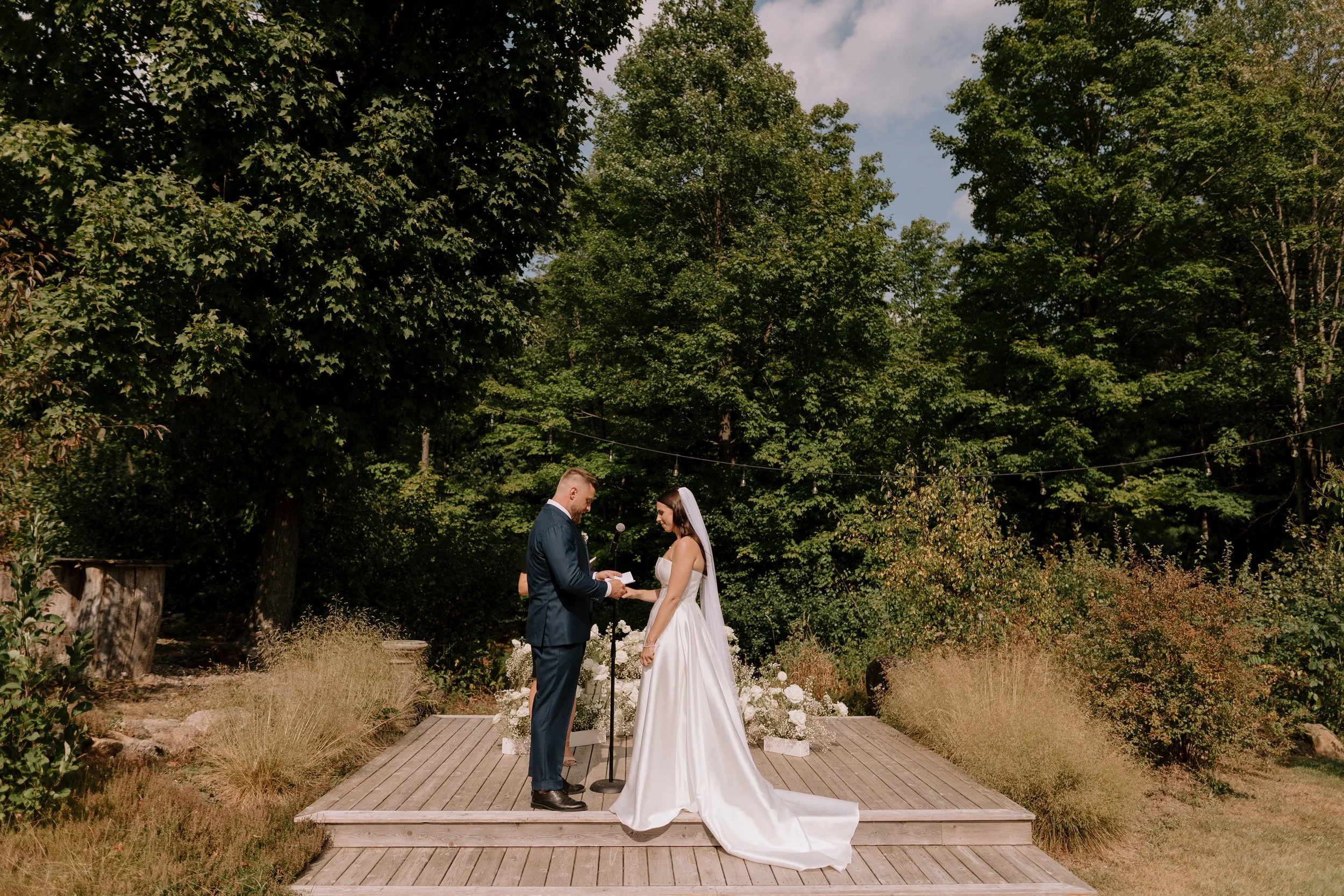 A bride and groom exchanging vows during an outdoor wedding ceremony beneath tall green trees on a wooden platform, with wedding flowers and decorations around them.