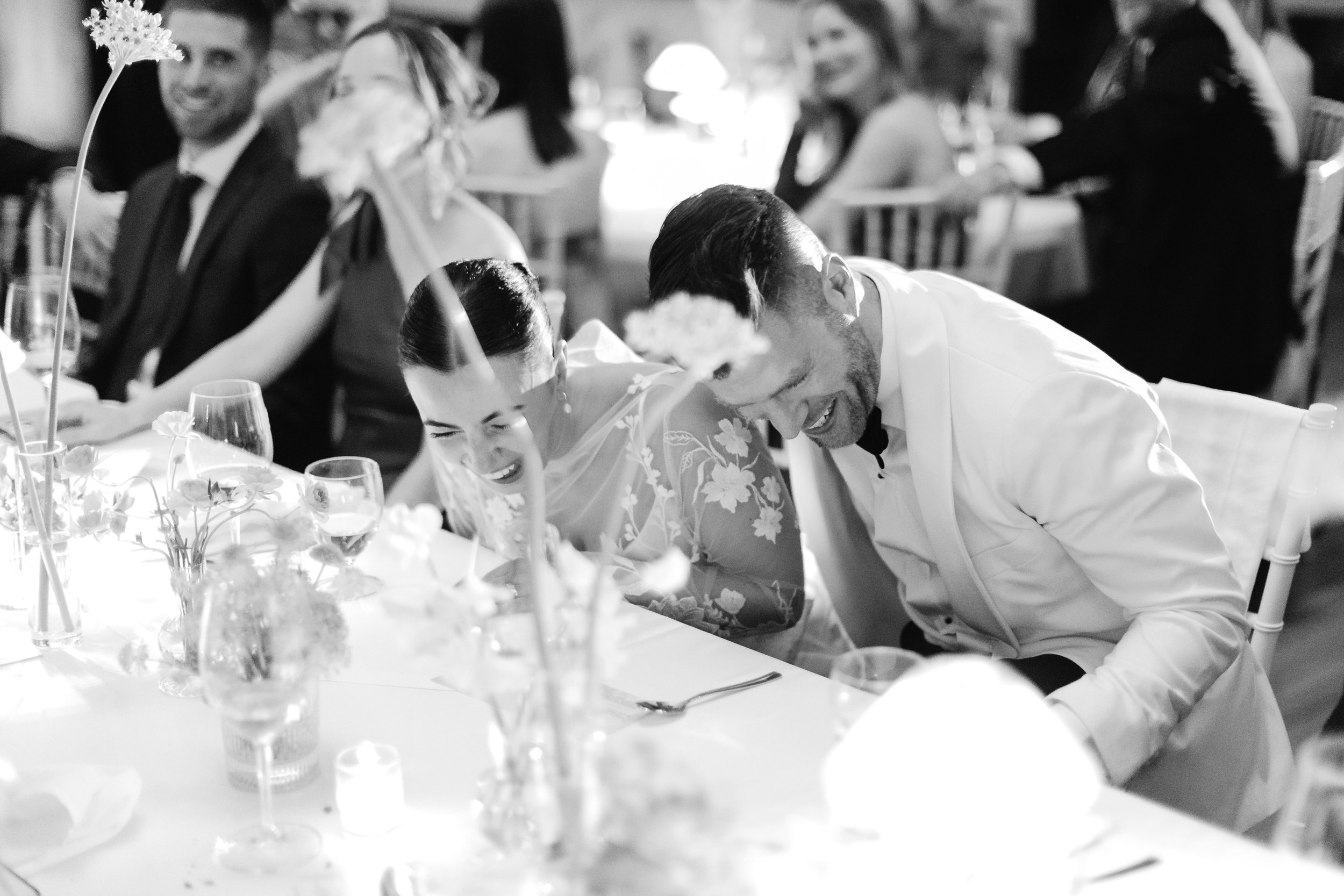 People at a wedding reception, sitting at a decorated table with flowers and glasses, laughing and enjoying themselves.