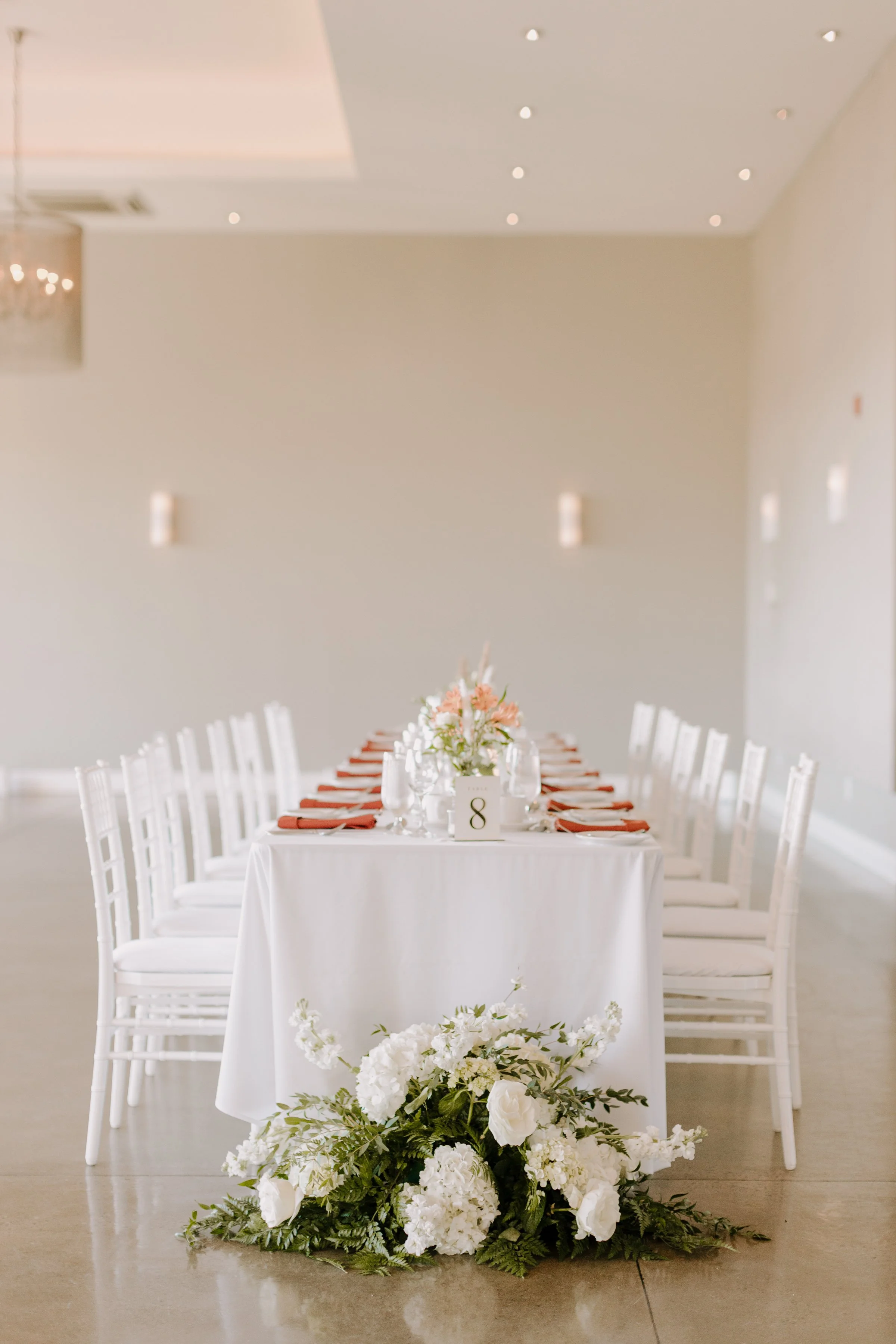 A decorated banquet table for a wedding reception, decorated with pink and white flowers, white chairs, white tablecloth, and place settings with white plates and pink napkins. A floral arrangement is at the center, and a table number '8' is displaye