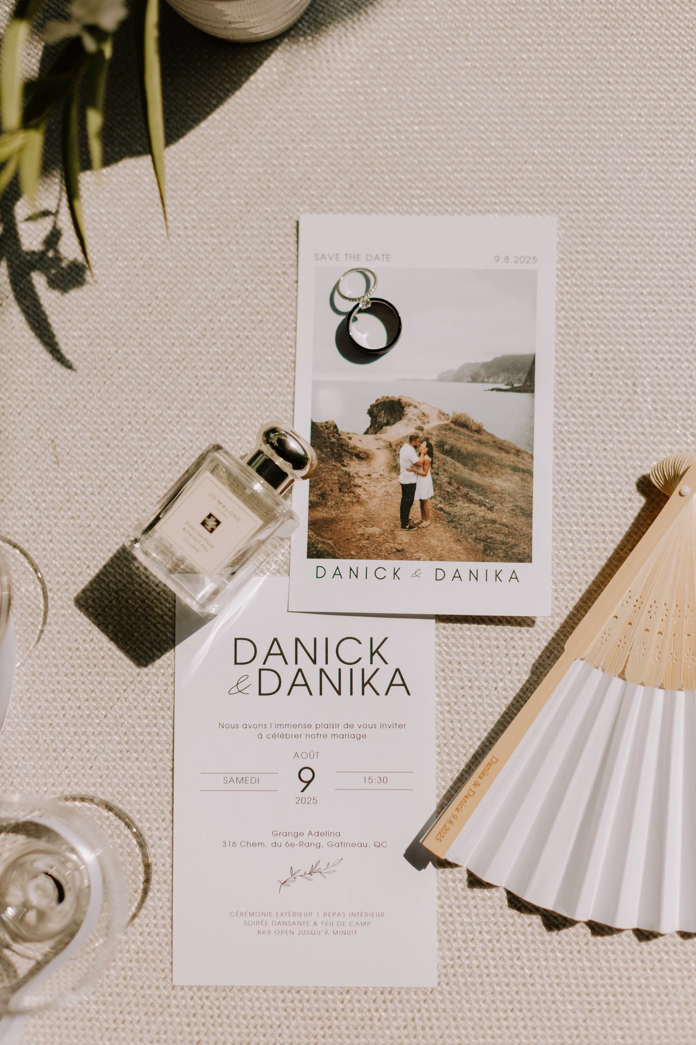 A wedding invitation with a photo of a couple by the sea, a 'save the date' note, perfume bottle, rings, and decorative items on a textured surface.