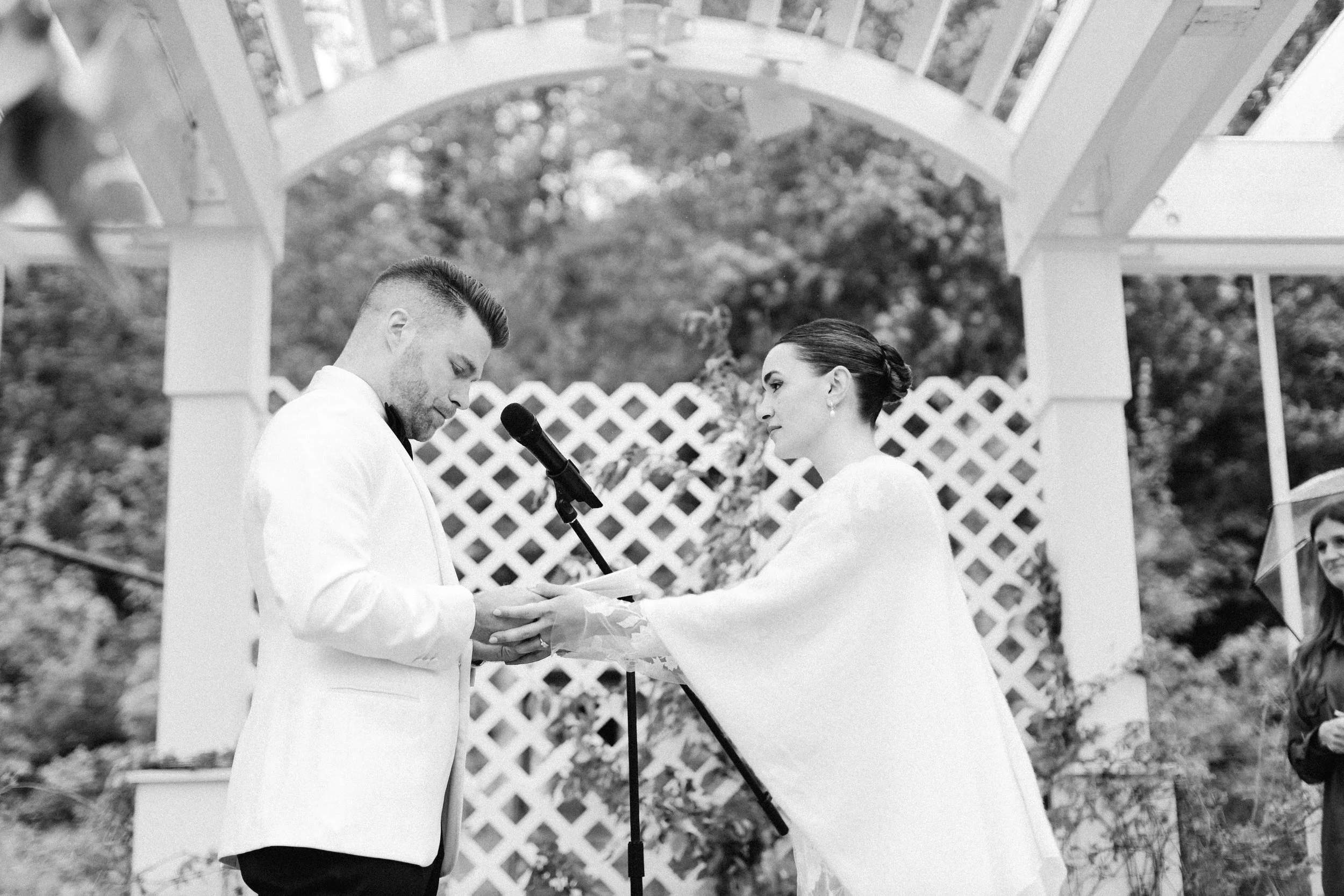 Black and white photo of a wedding ceremony with a man and woman holding hands, standing under a wooden arch, exchanging vows. The officiant is reading from a paper, and a person is sitting nearby. The background features trees and a white lattice fe