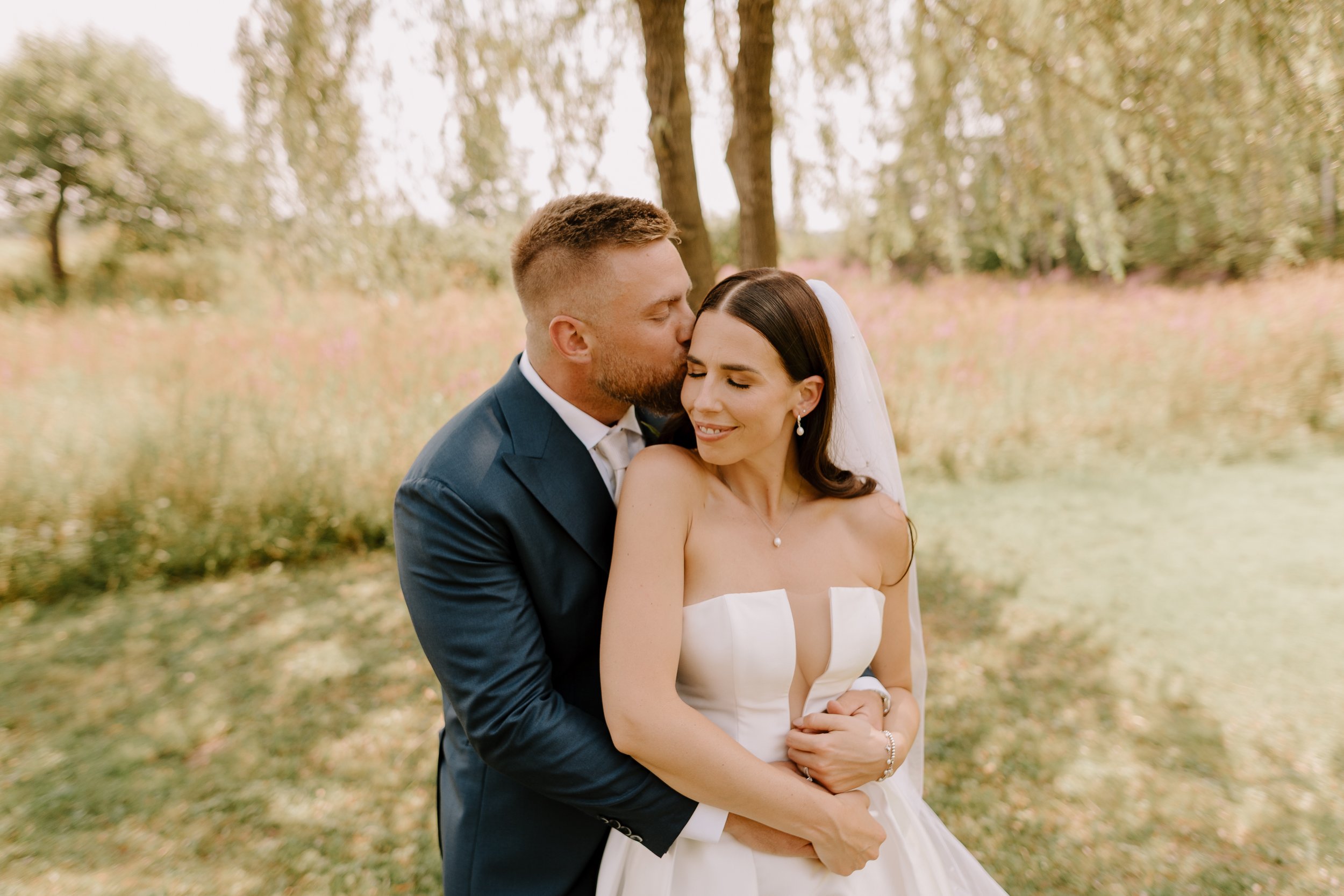 A bride and groom share an affectionate moment outdoors. The groom kisses the bride on her temple as she smiles softly with her eyes closed. The background features trees and a natural setting.