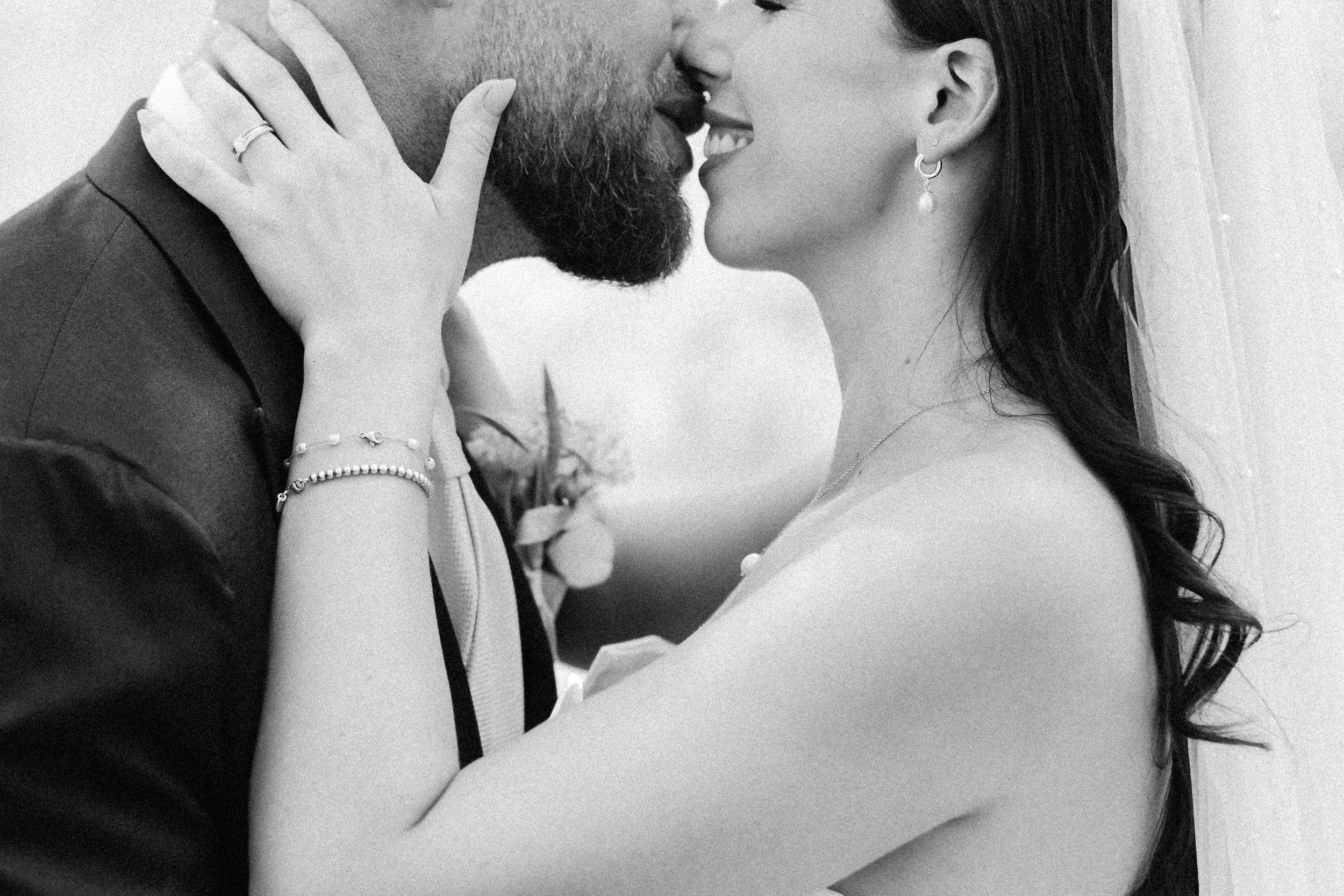 A couple is about to kiss, with their faces close together and eyes closed. The woman has long, dark, wavy hair and the man has a beard. The woman is wearing earrings, a necklace, and a bracelet. The man is wearing a dark shirt.